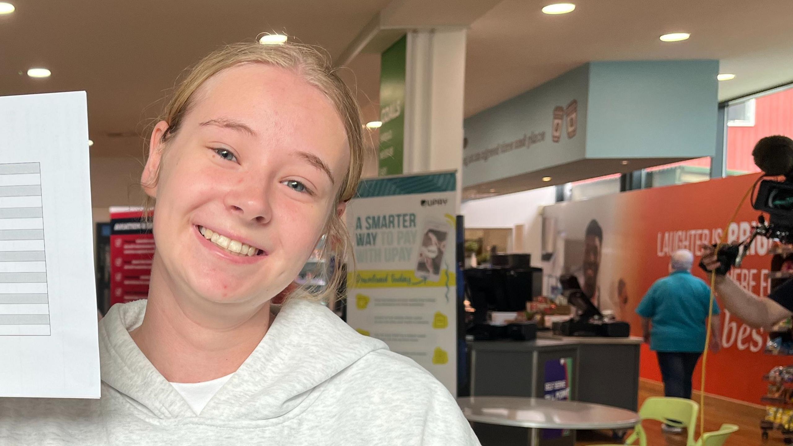 Olivia Melville poses smiling at the camera, wearing a grey hoodie, holding the paper with her results on just off the left of the screen.