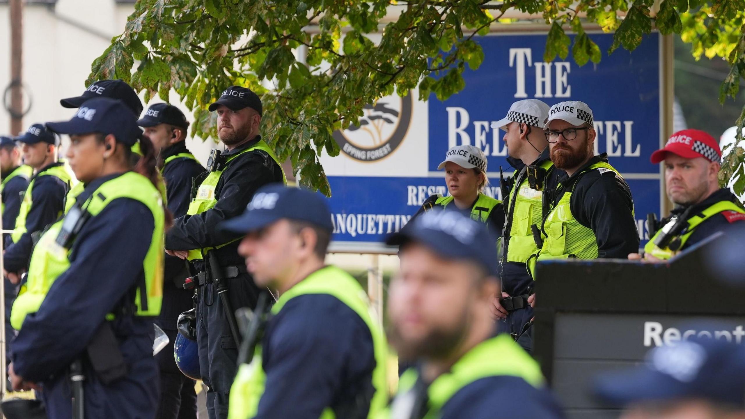A large group of police officers, all wearing caps and hi-vis vests, standing in front of a large blue sign outside The Bell Hotel, which advertises its name.