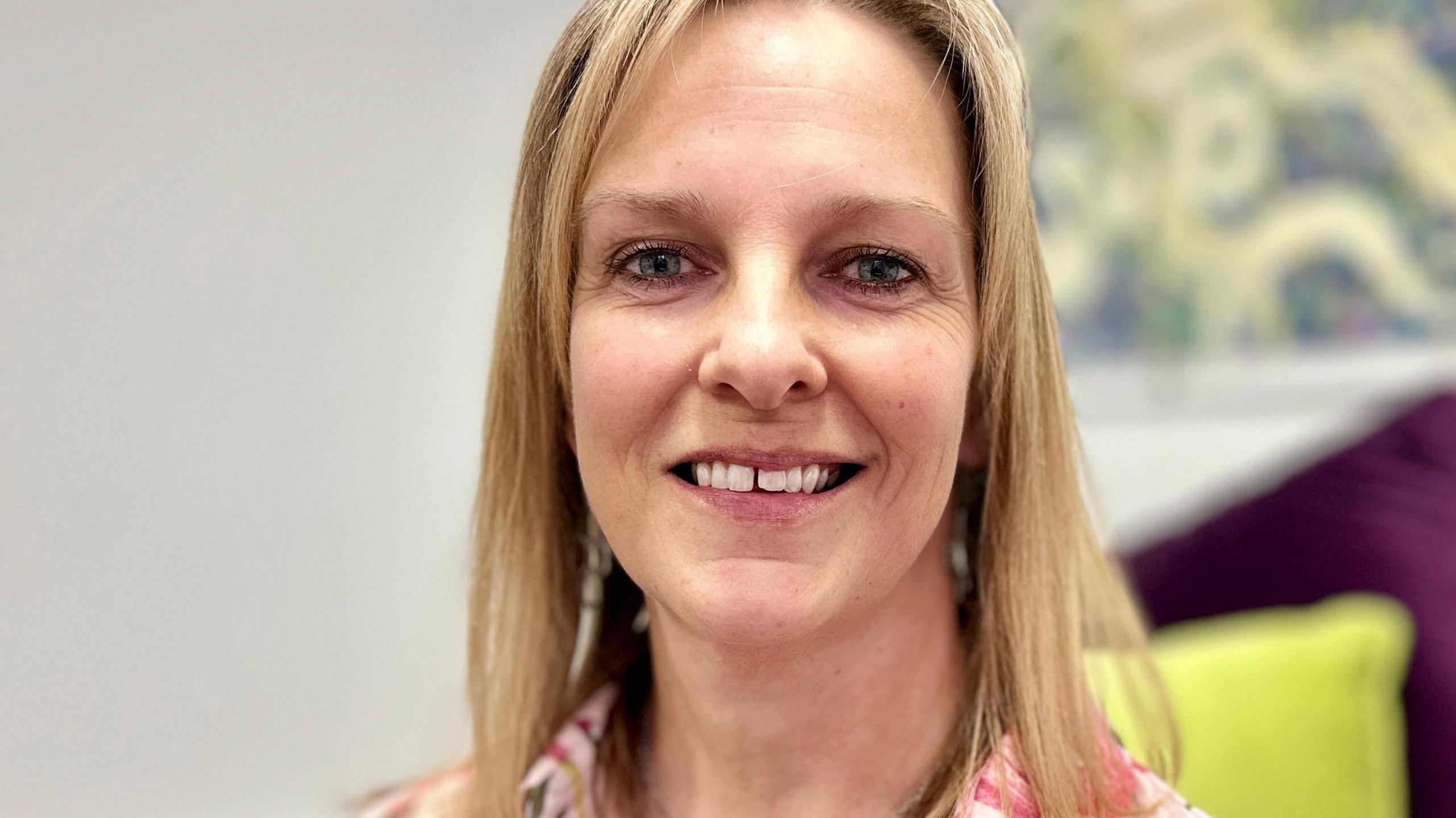 Portrait photograph of Becky Ward smiling to camera. She has long blonde straight hair and is wearing a floral blouse.