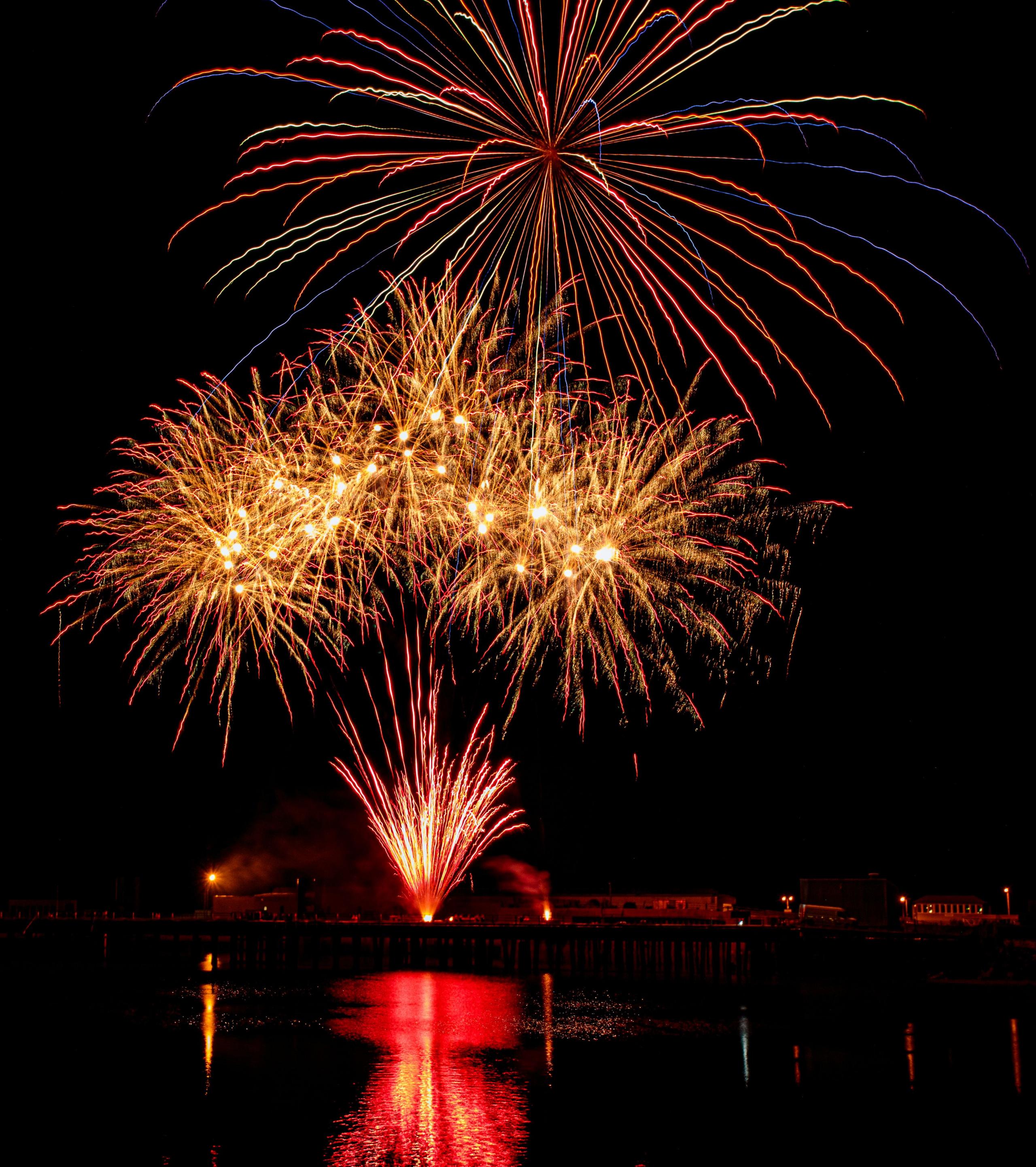 Fireworks in red and orange and yellow light up a dark sky and are reflected in the water