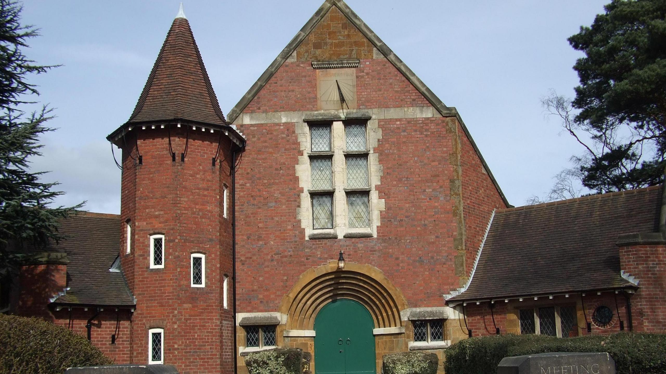 A large brick building with a three-storey tower next to it. Both buildings are created from red brick and the tower has a pointed roof. Behind them is blue sky.