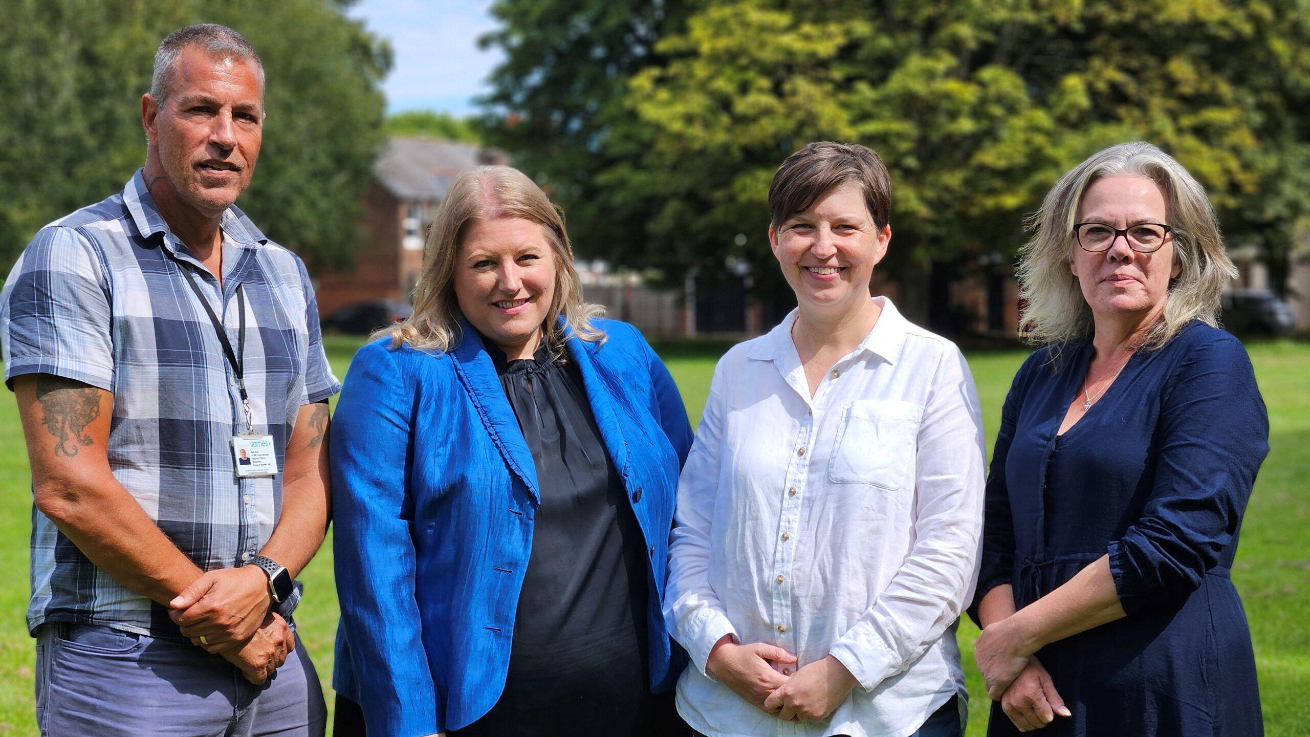 A grey-haired man in a checked shirt and blue chinos, a blonde woman in a navy dress and a royal blue blazer, a short-haired woman in a white shirt and blue trousers and a grey-haired woman with glasses and wearing a navy dress stand on a grassy area with trees behind them. 