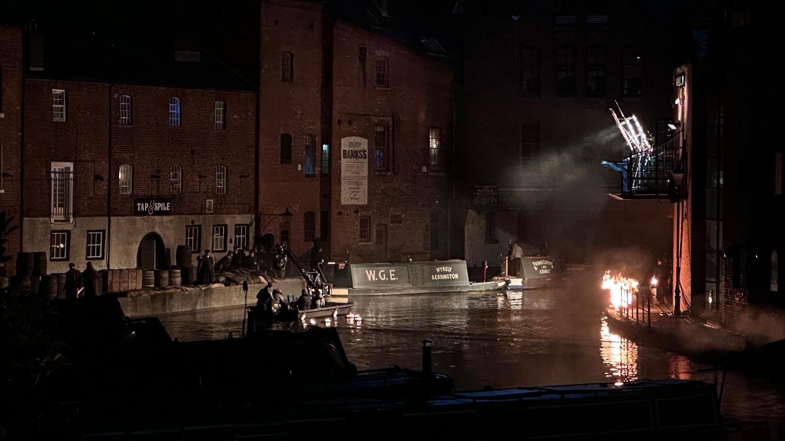 A canal at night with redbrick warehouse-style buildings on either side. There are large professional studio lights installed on a balcony and pointing at a group standing on a towpath. A fire has also been created on one side of the canal.