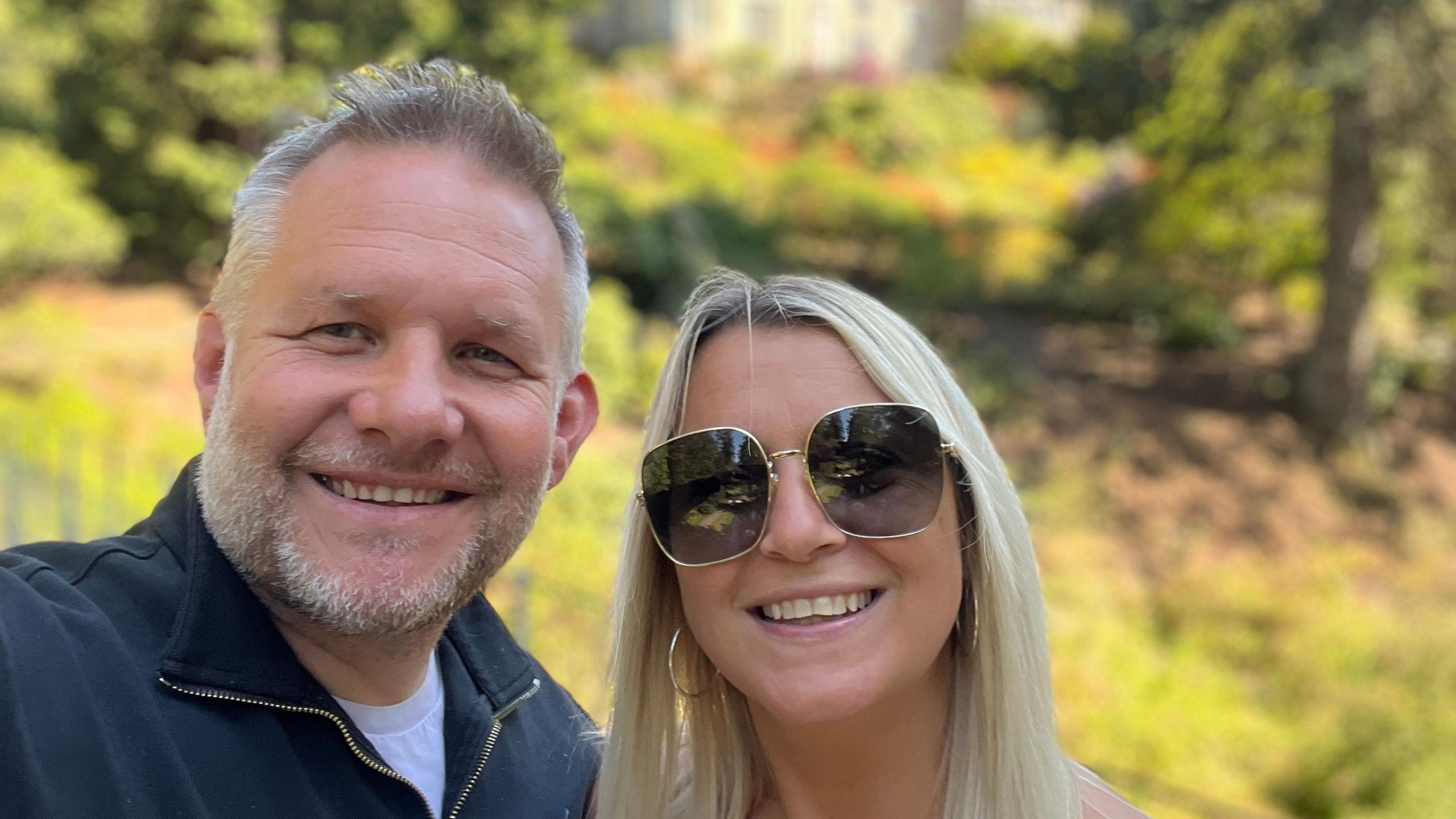 Selfie photograph of Ben Giles (left) and his wife Lindsey(right). Ben has grey short hair, a grey beard, and blue eyes. She smiles widely at the camera. Lindsey wears big wide sunglasses with a gold frame, gold hoop earrings and smiles at the camera. She has bright blonde long hair. Behind them is green grass highlighted by the sun, some trees and a manor house.