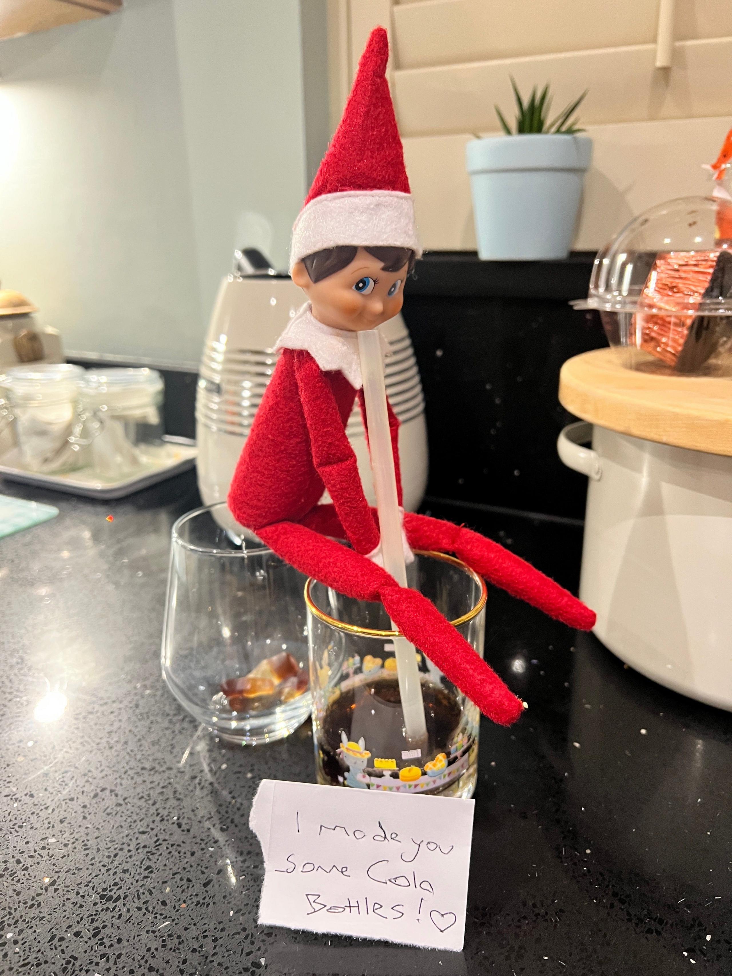 An elf sat on a glass drinking cola. The second glass has cola bottle sweets in underneath its bottom.