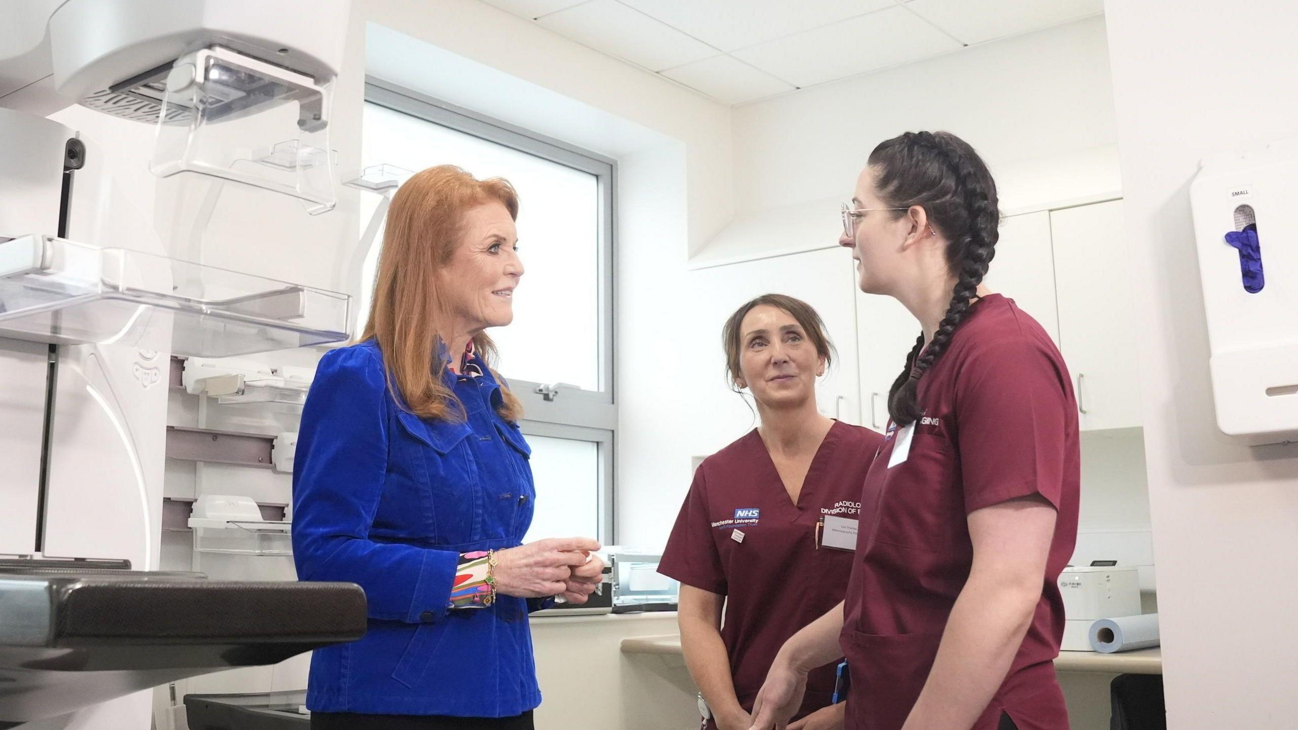 Wearing a royal blue blazer, the Duchess of York speaks with two staff in maroon scrubs during a visit to Prevent Breast Cancer's headquarters in Manchester. They are photographed in a lab at Wythenshawe Hospital's Nightingale Centre.