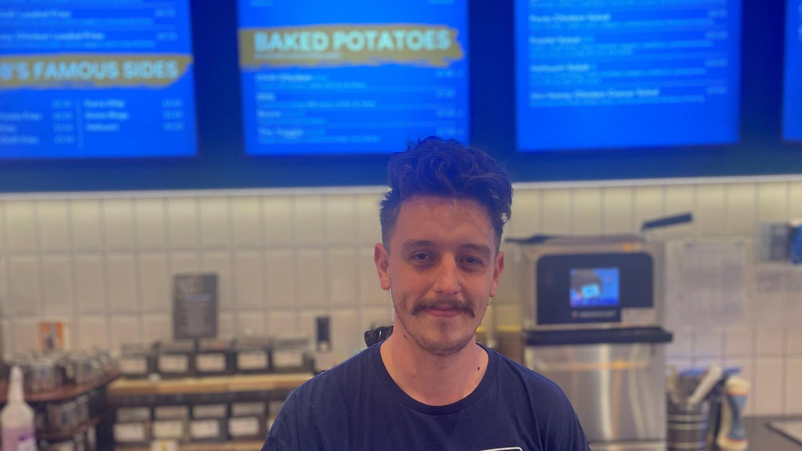 A man wearing a navy T-shirt stand behind a cash register - behind his head menus can be seen and a small silver oven