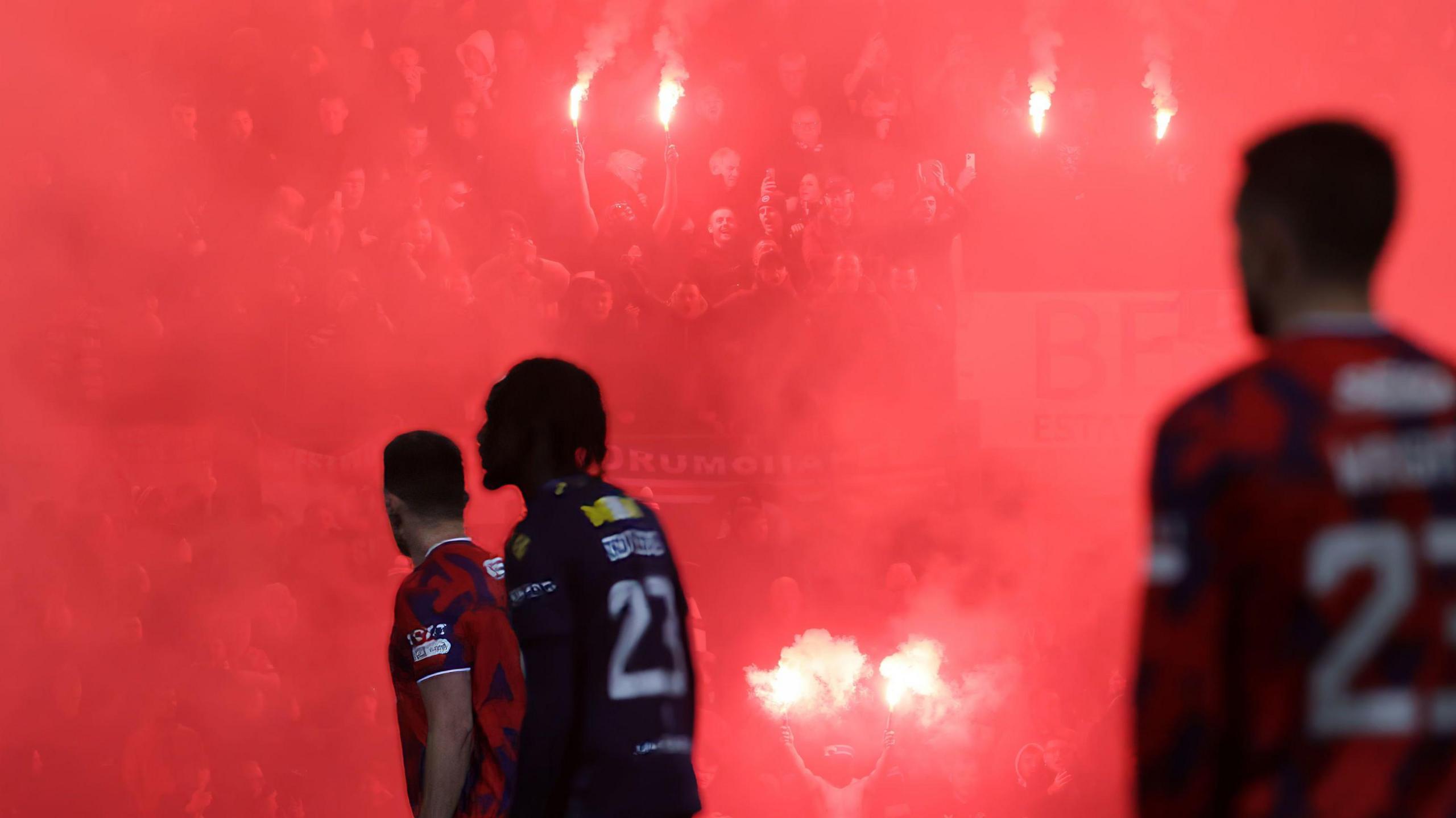 Players from Dundee and Rangers in the foreground as Rangers fans hold red flares in the background