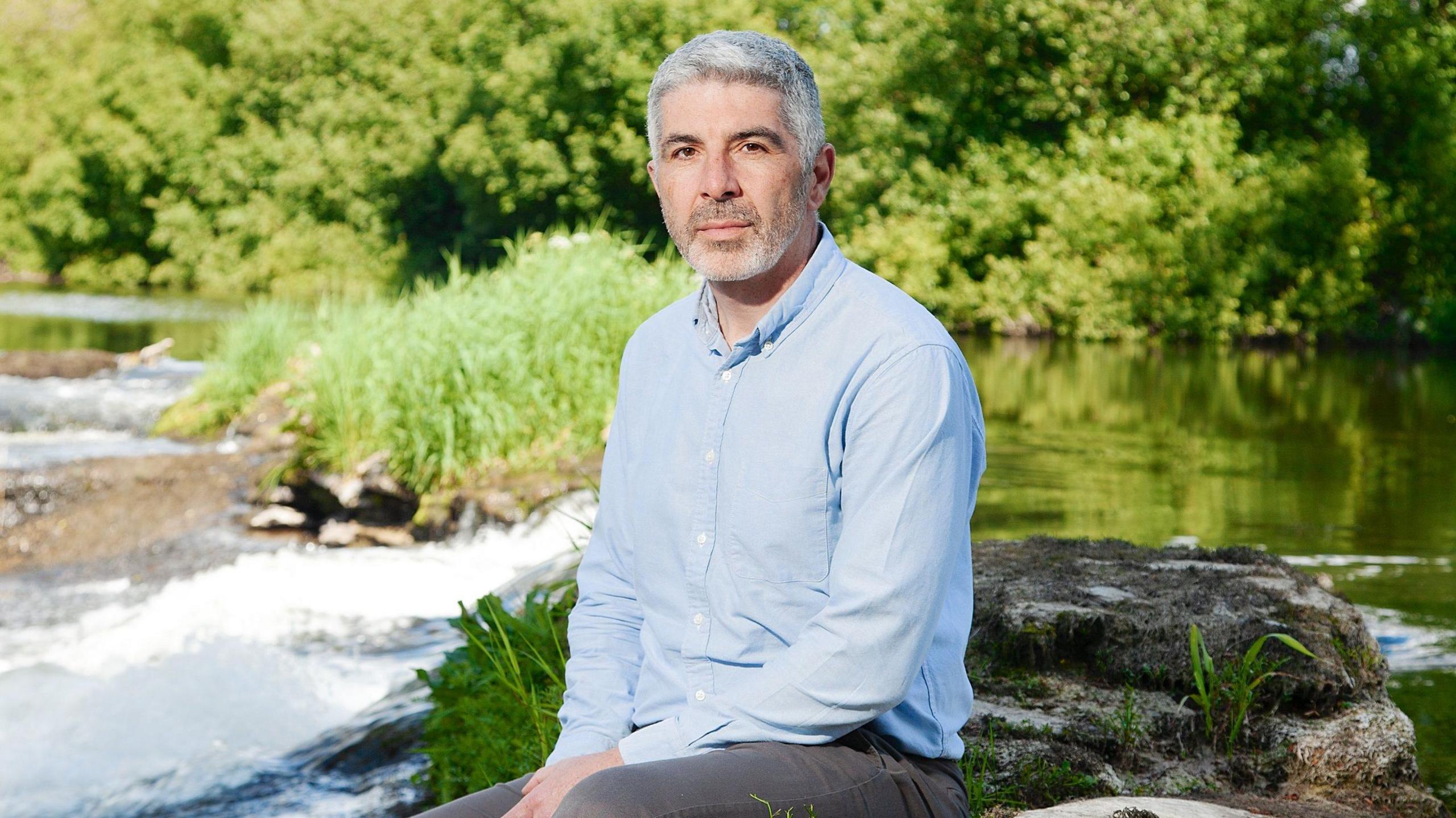 Derek Walker, Wales' Future Generations Commissioner sits on a rock with a river in the background. He's wearing a blue shirt and black trousers.