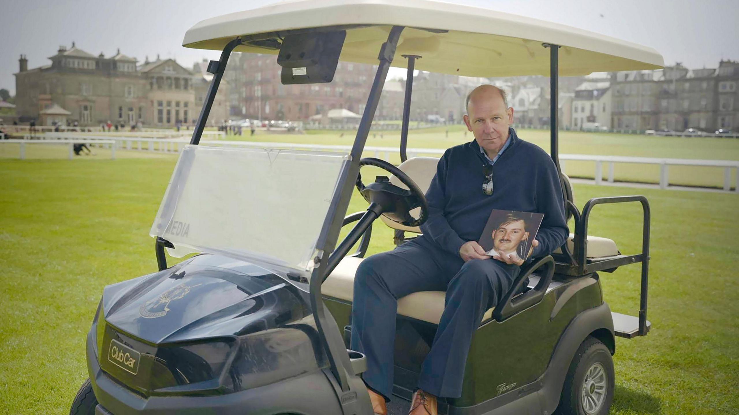 Stewart Smith is wearing a blue jumper and trousers and is sitting in a blue gold buggy beside the first hole at St Andrews' Old Course. The Royal and Ancient building is in the background.