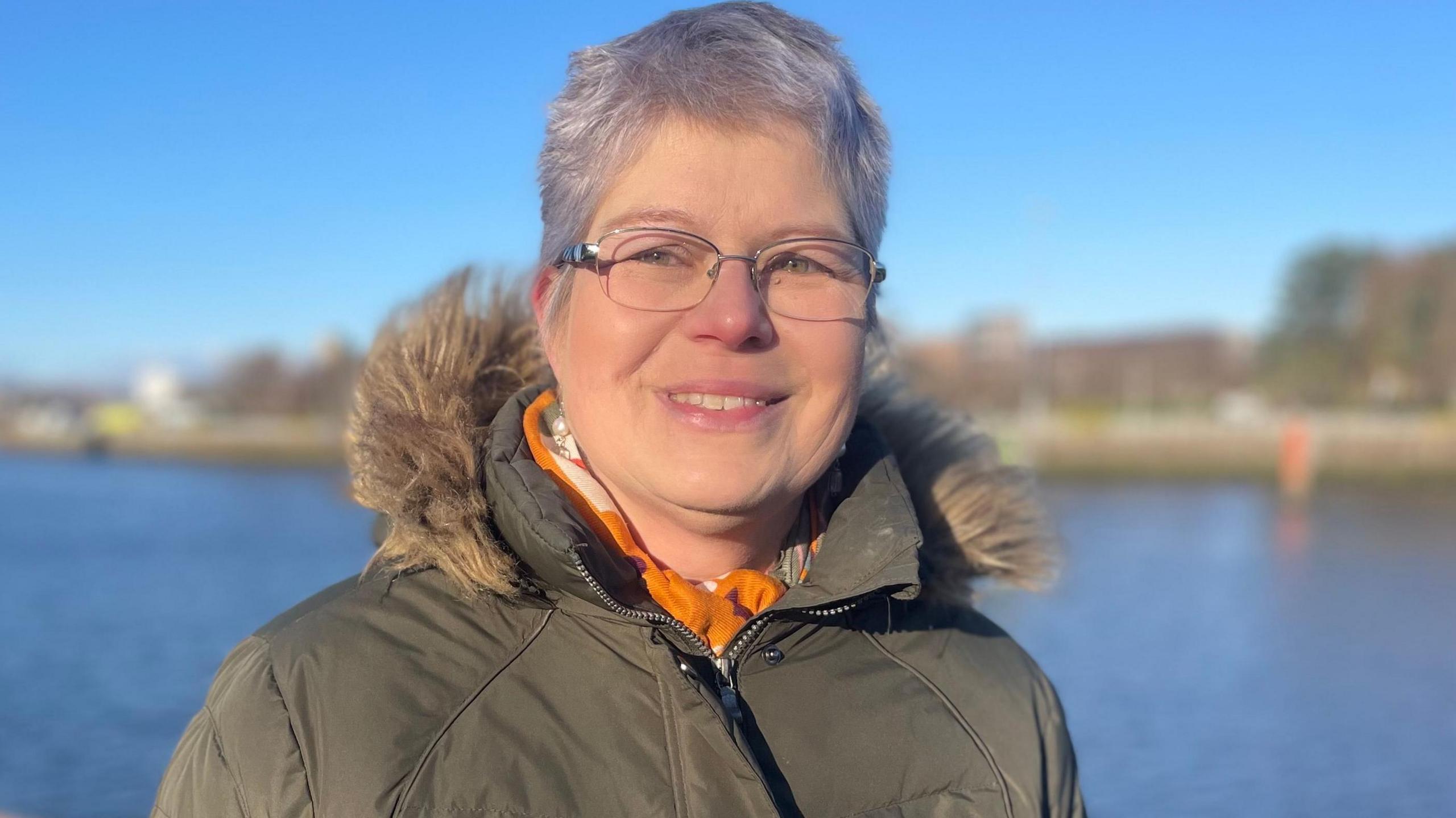 Steffi is standing outside by a stretch of water looking directly at the camera. It appears to be cold day and she is wearing a greenish winter coat with a furry hood. She has short grey hair and rimless glasses. 