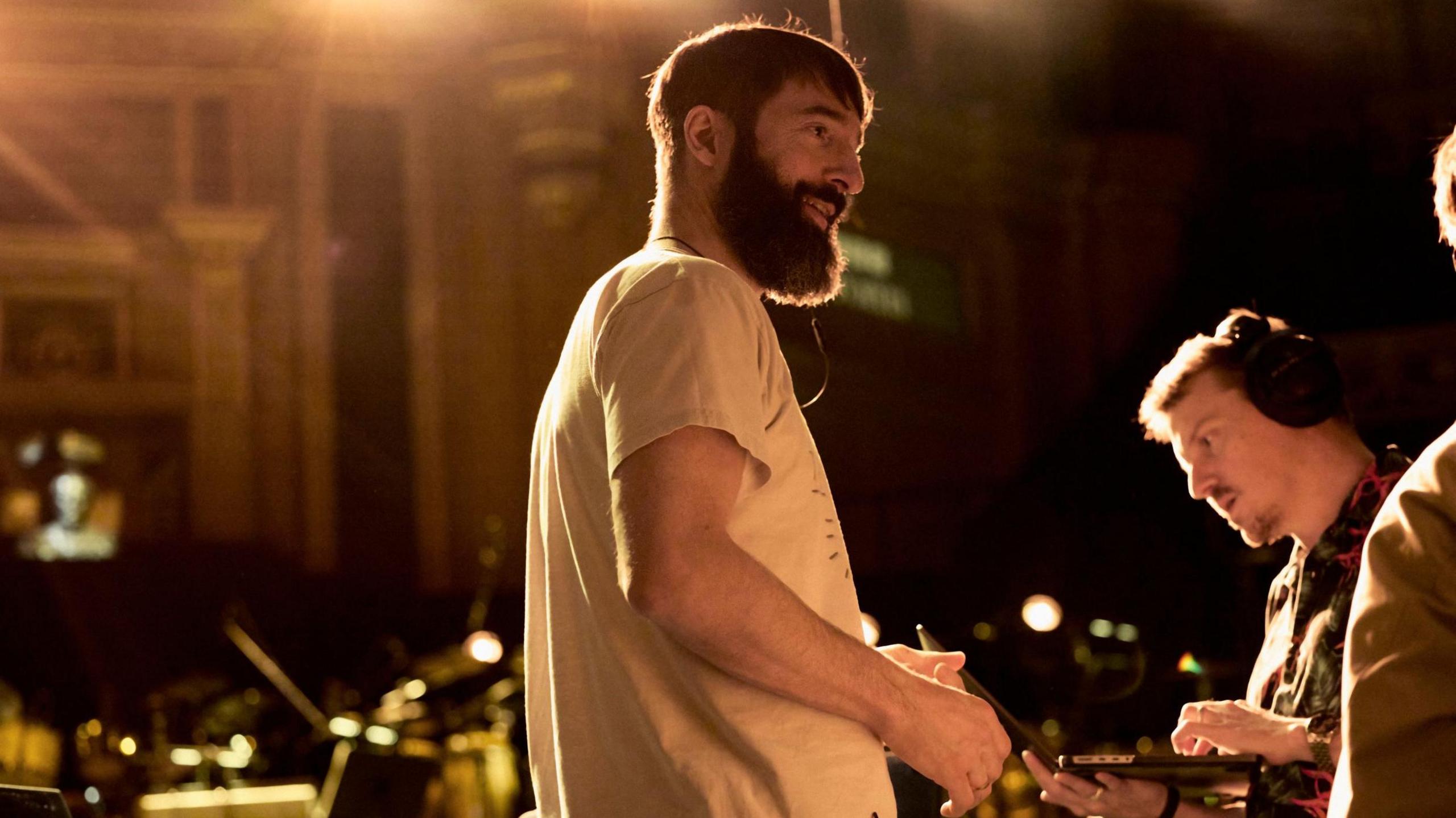 Jules stands sideways on, smiling and talking to people on his right, one of whom is wearing black headphones. They appear to be at the Albert Hall with professional lighting shining. Jules wears a white t-shirt and has a thick beard and moustache and short smart brown hair.