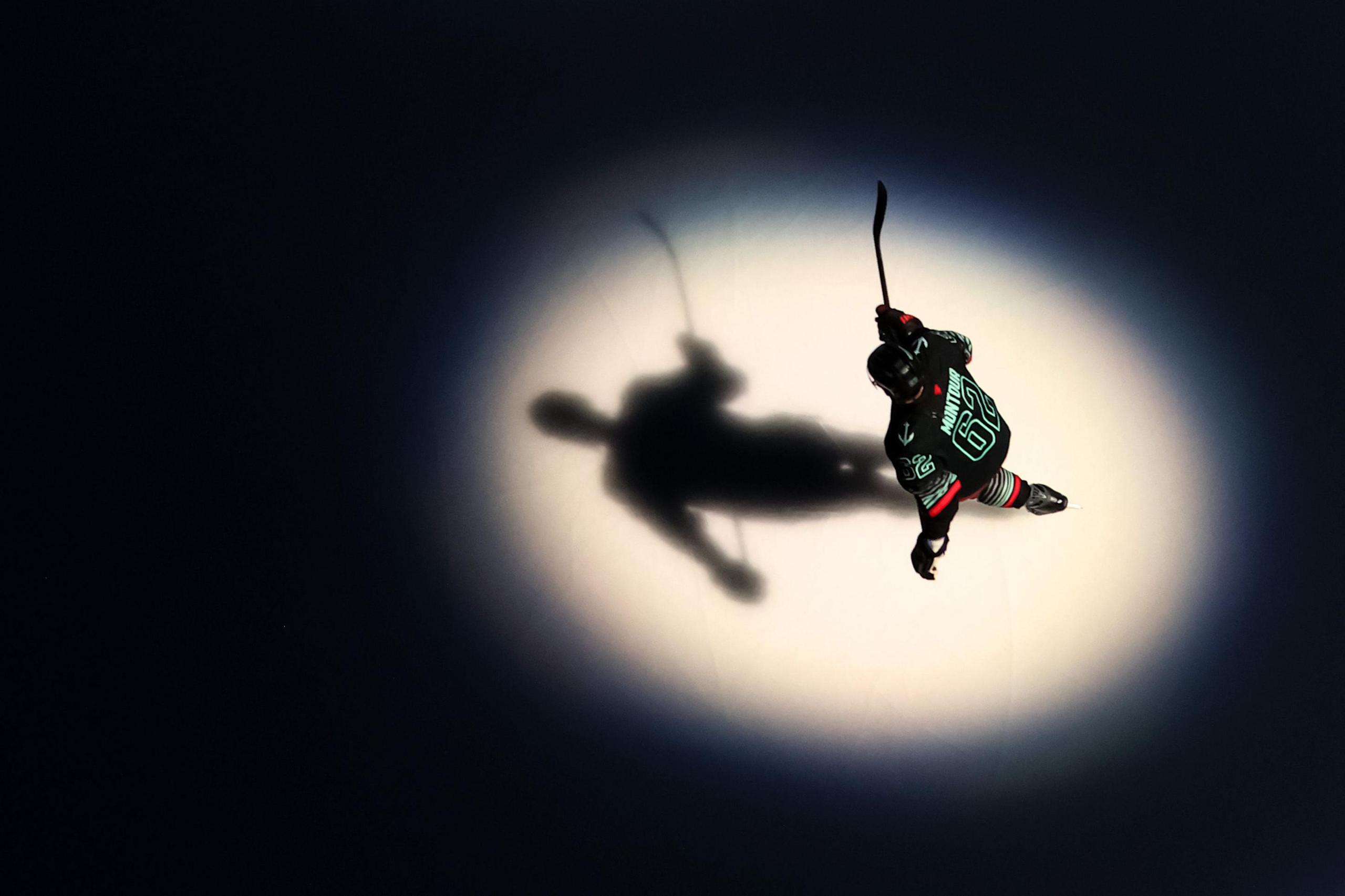 Un jugador de hockey patina solo bajo un foco brillante sobre el hielo oscuro, proyectando una sombra nítida debajo de él mientras sostiene su palo levantado a mitad de la zancada.