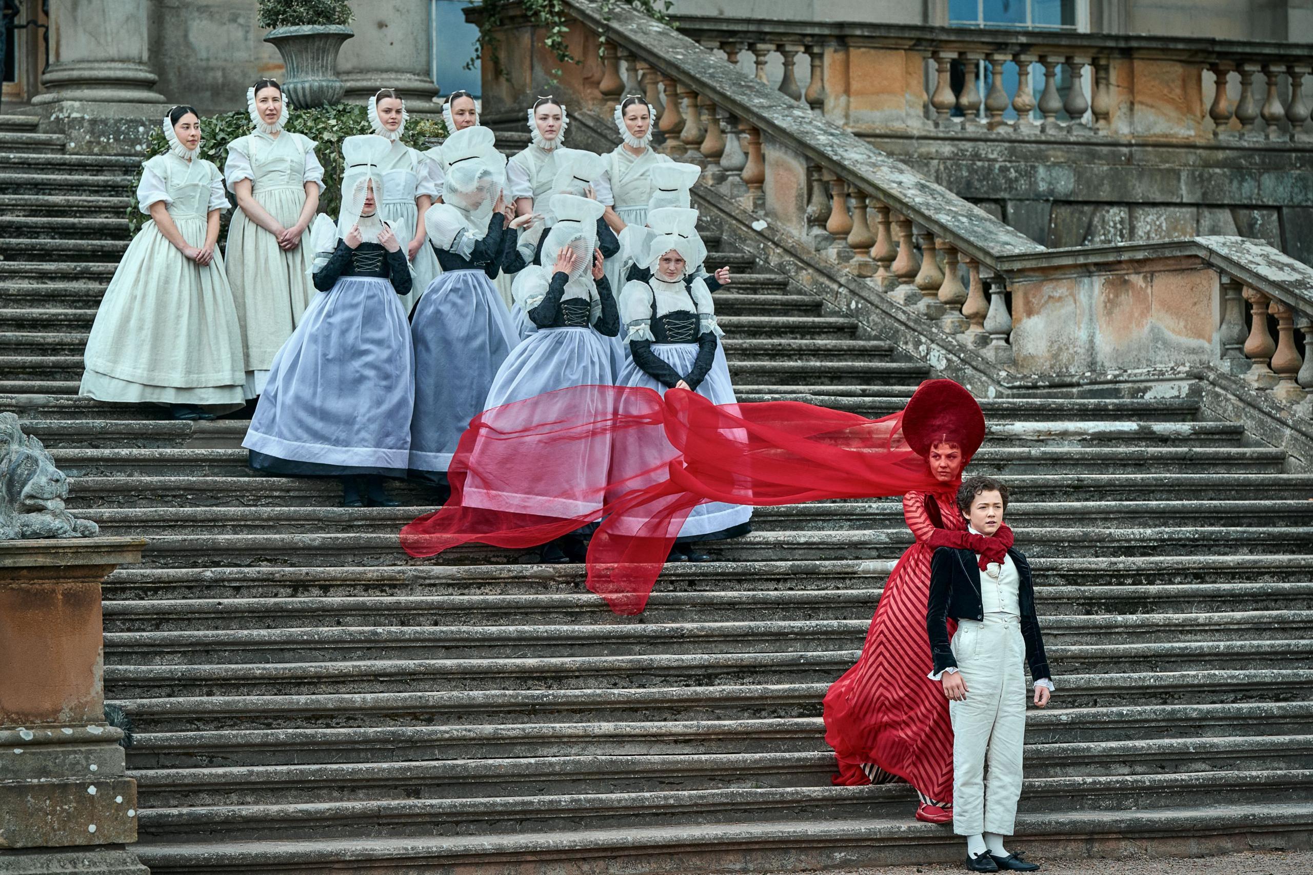A long stone staircase with a a group of ten women in period domestic costume standing in a group halfway up. In front of them a woman stands in a bright red, elaborate flowing dress. She has her arms around a young man, both facing front. He is wearing formal clothes of a short, dark jacket, white shirt and waistcoat and white trousers. 