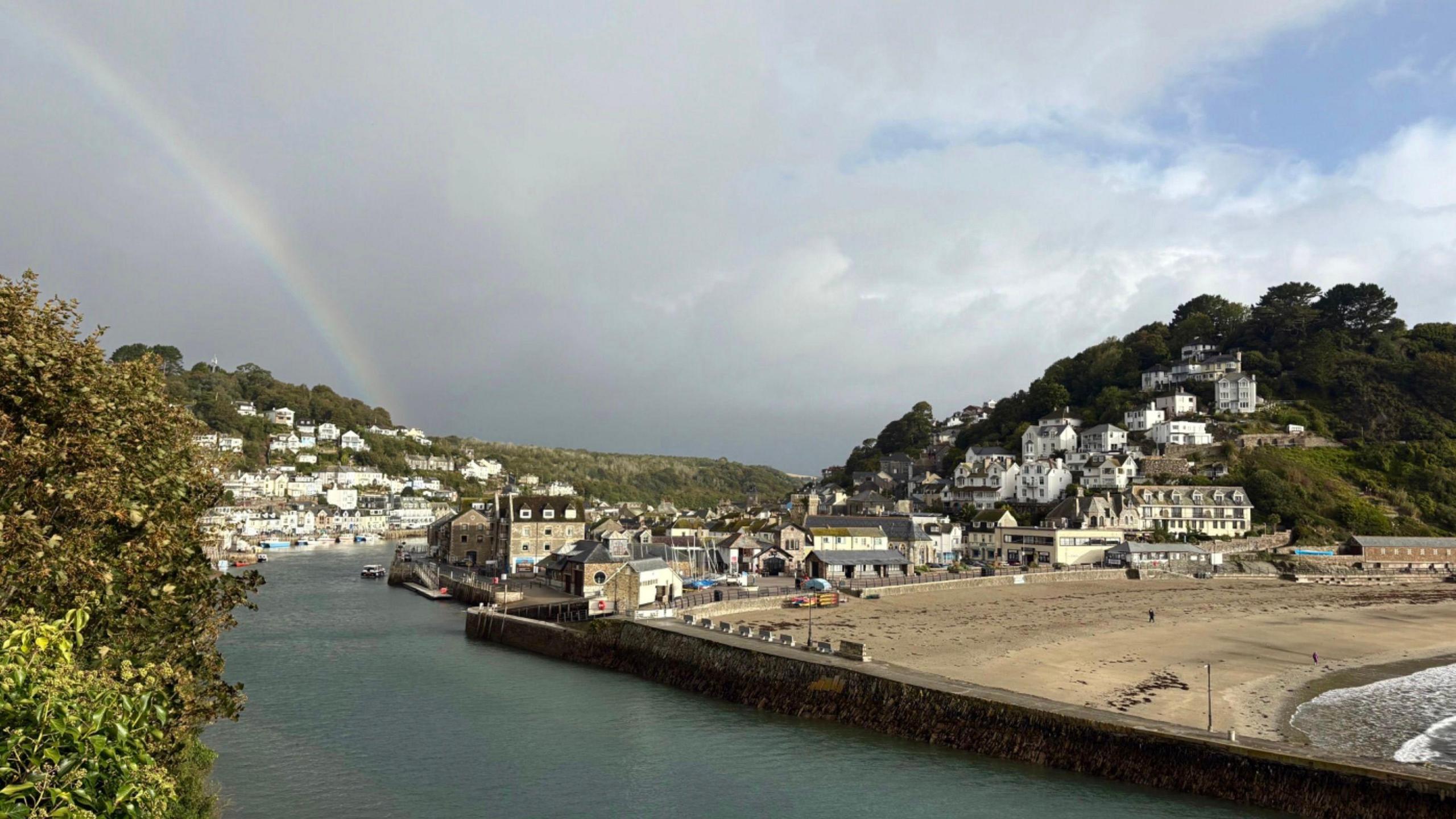 Looe is seen slightly in the distance. A number of shops and boats are pictured. There is a beach to the right, and a rainbow in the distance.