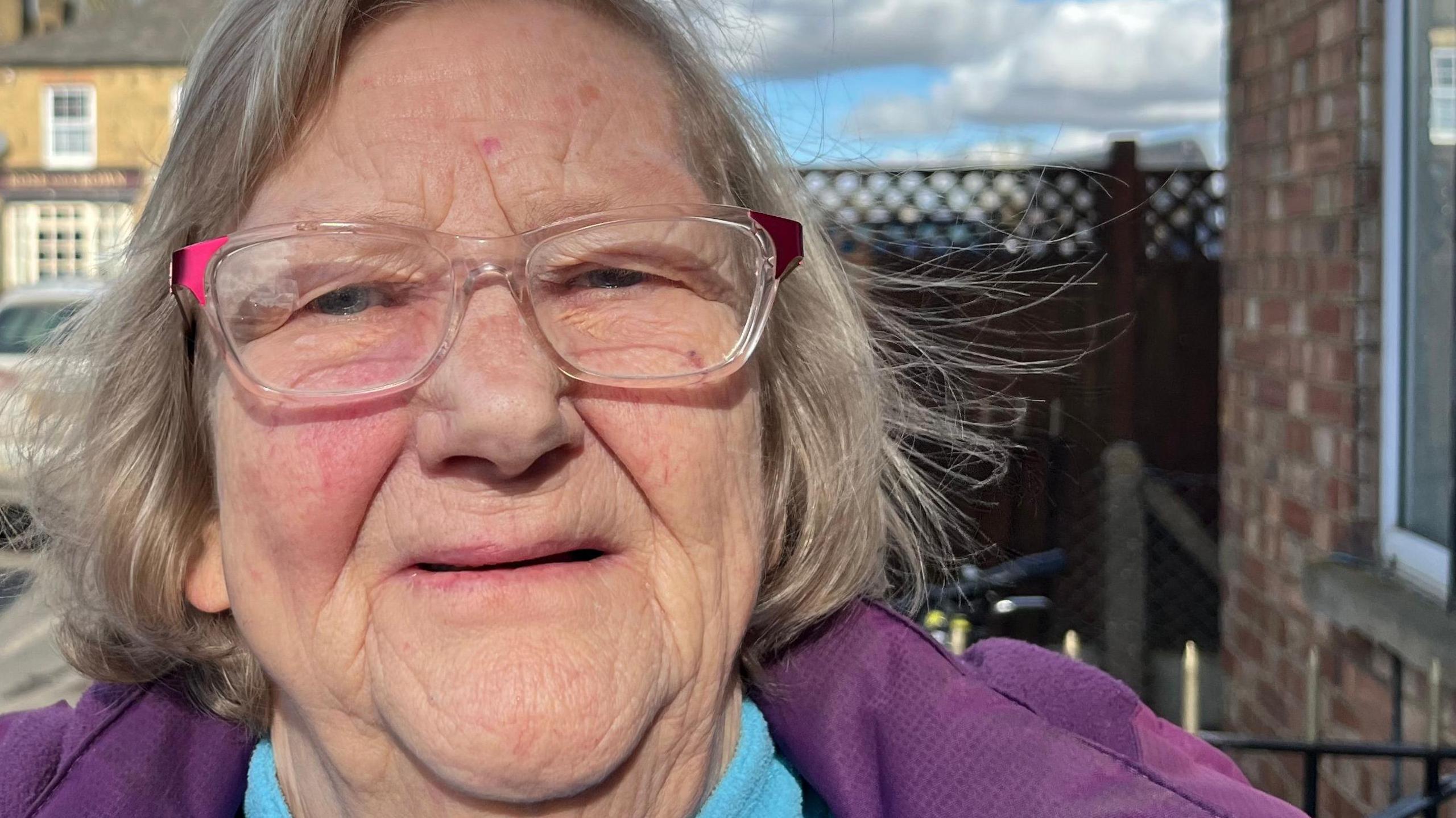 Valerie Shadbolt, 68 has sandy coloured hair parted to the left she is wearing glasses with pinkish tints to the frames she has a purple coat on over a sky blue fleece top, behind her is a parked minibus and car and some housing can be seen too.