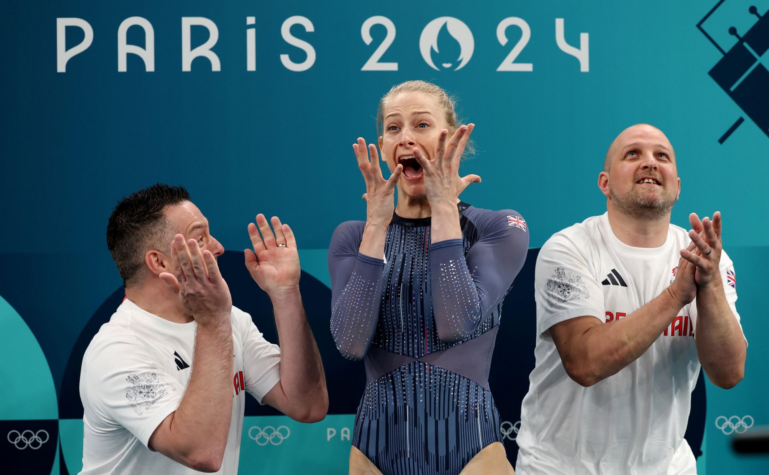 Bryony Page, in a blue sparkly leotard, reacts in shock with her hands raised to her face and her mouth open in surpise. She is stood between her coaches, two men in white t-shirts, who are clapping.