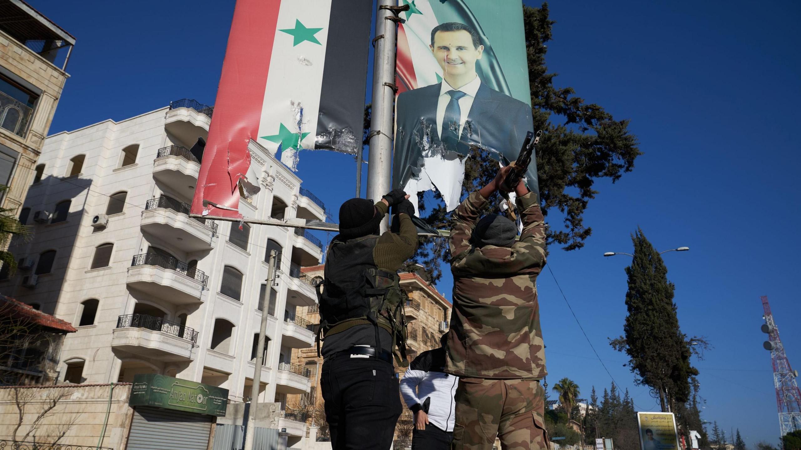 The backs of two men tearing down a banner of Syria's President Bashar al-Assad. They are wearing camouflage.