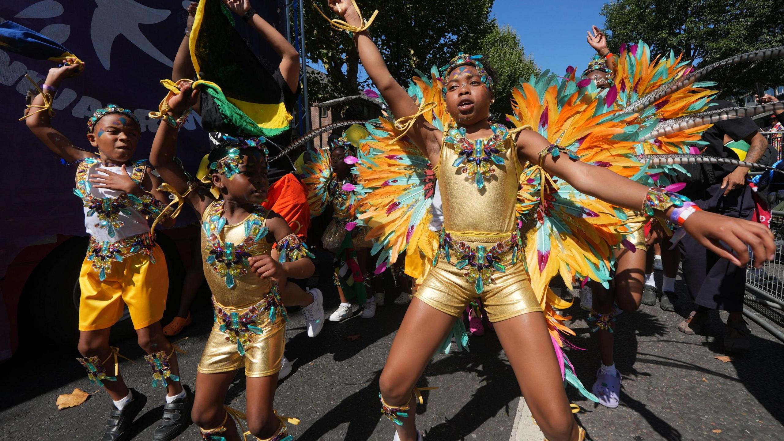 A group of children in gold costumes with orange and teal feathers dance down the street.