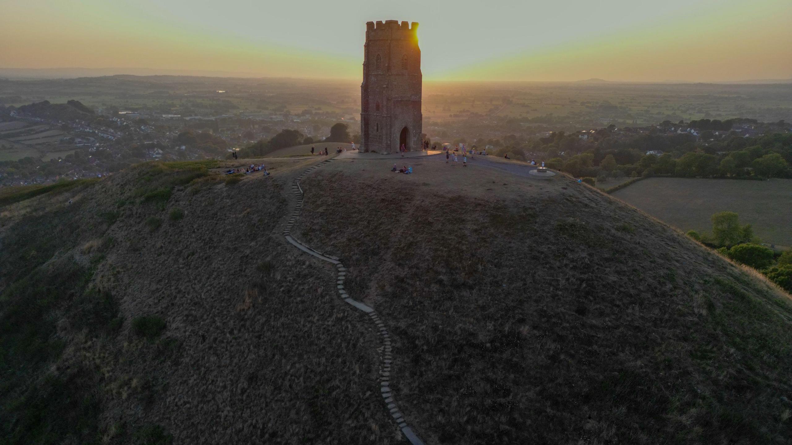 A shot of Glastonbury Tor. There is a steep hill with a rectangular shaped tower on top of it. The landscape features miles of fields and trees. The sun is setting in the sky.