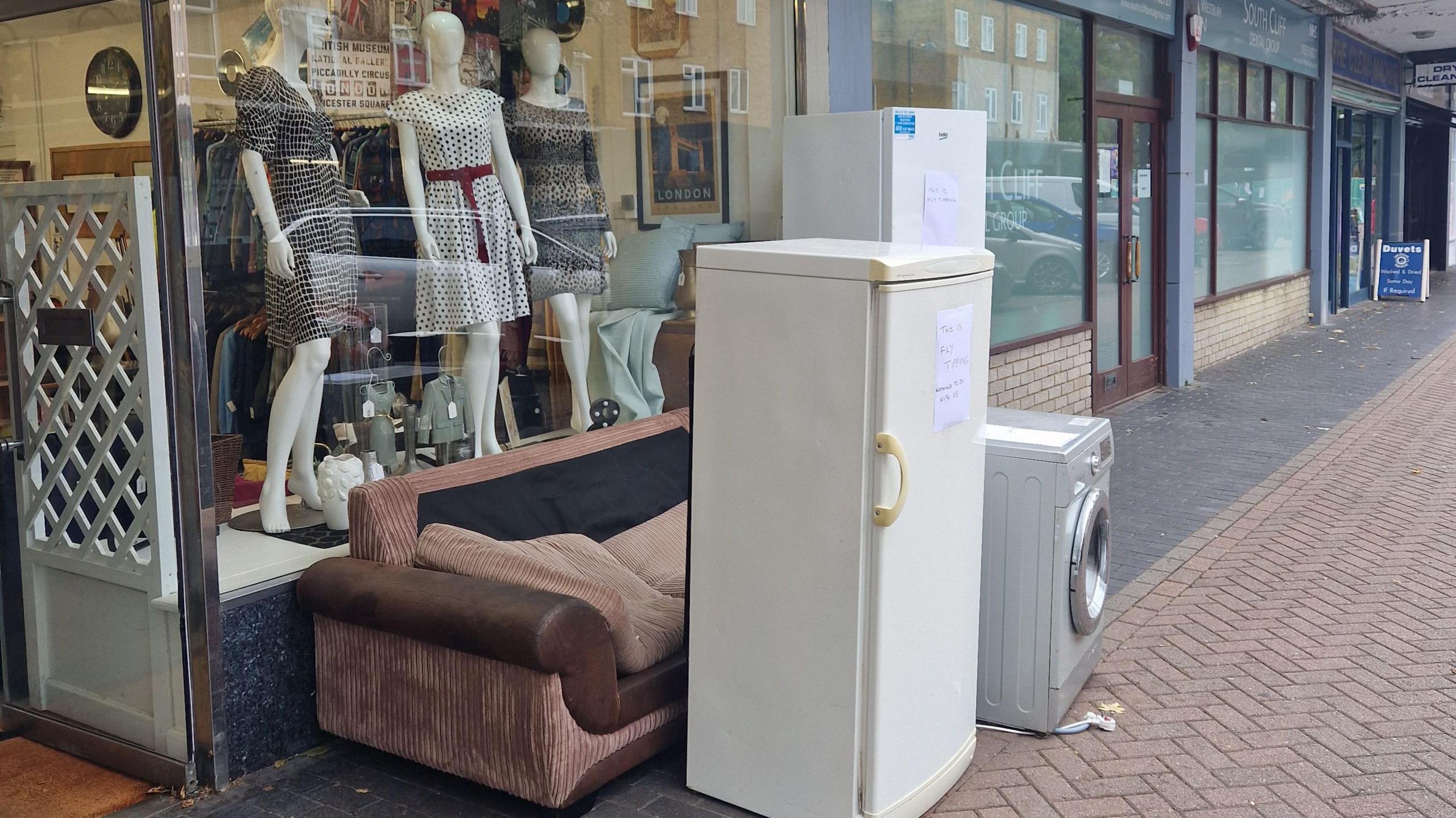 A sofa, two fridges and a washing machine outside the Imperial Charity shop. The road surface is made of interlocking bricks and other shop units are visible alongside the charity shop
