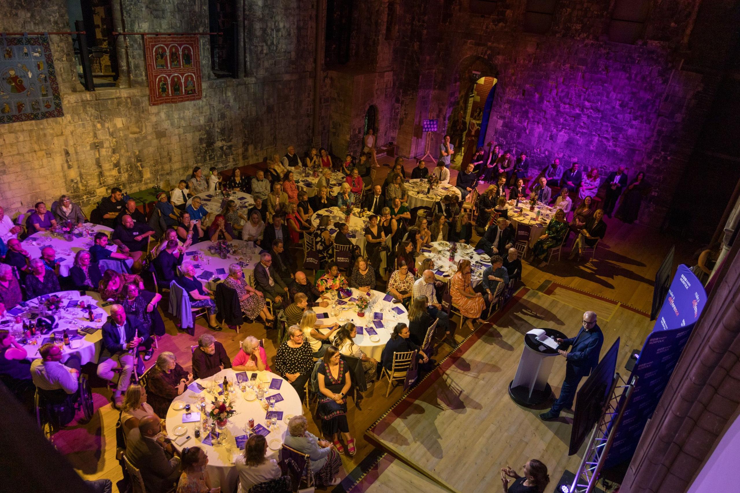 Eleven round tables are on a wooden floor, each has people sitting around them drinking and eating. The lighting is white and purple coloured. The walls of the room are old flint.