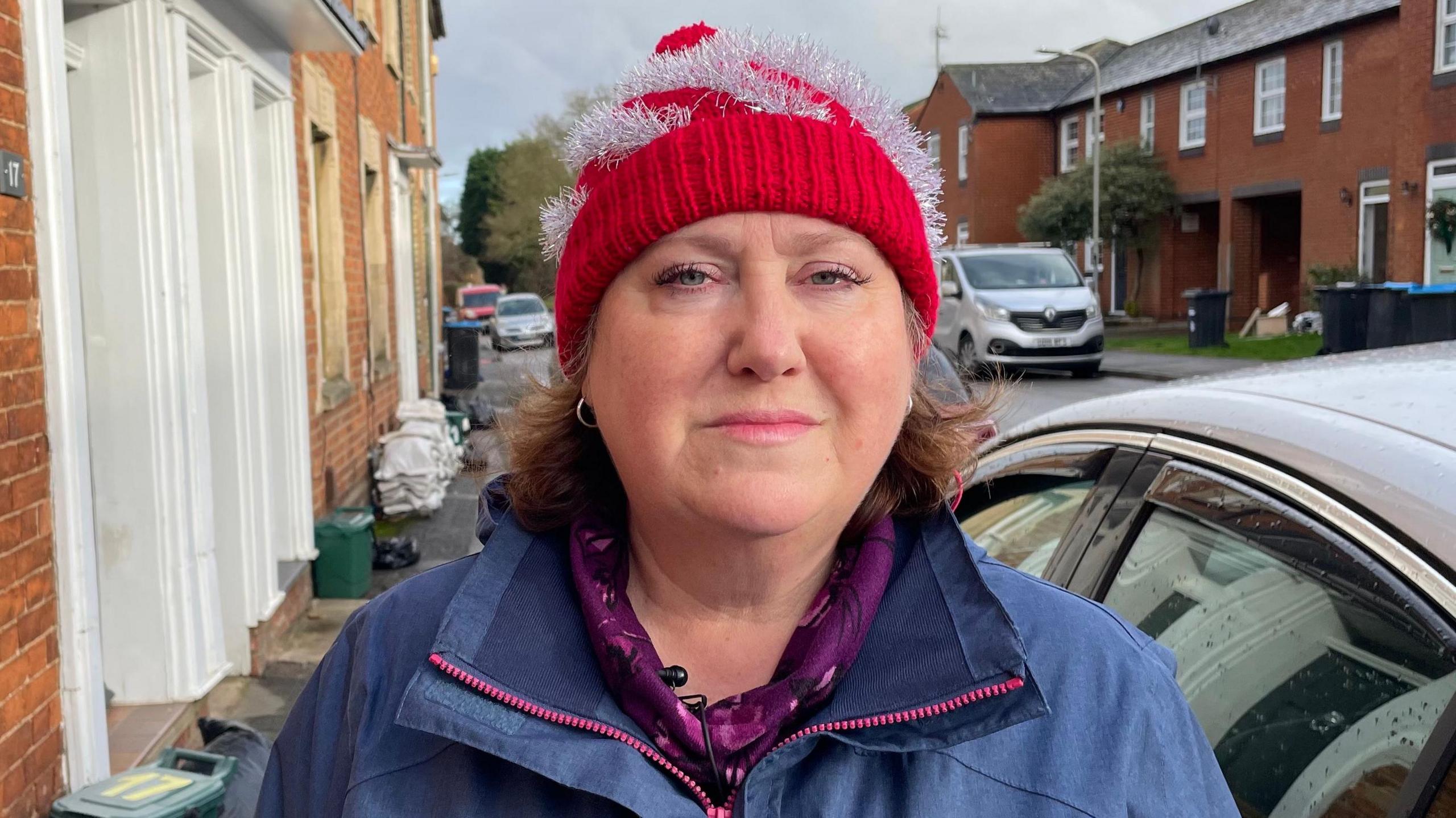 Jane Carr, looking direct to camera and standing on the pavement of a residential street. Jane is wearing a red and silver festive bobble hat and a blue waterproof coat, with a purple coloured top underneath. She has blue eyes and her light brown hair can be seen emerging from under the red hat. Immediately to the right of her in the image is the side of a car. A silver van is parked further away behind her on the opposite side of the road. To the left of the image are the frontages of terraced houses. On the pavement are some small foodwaste bins, as well as a pile of sandbags.