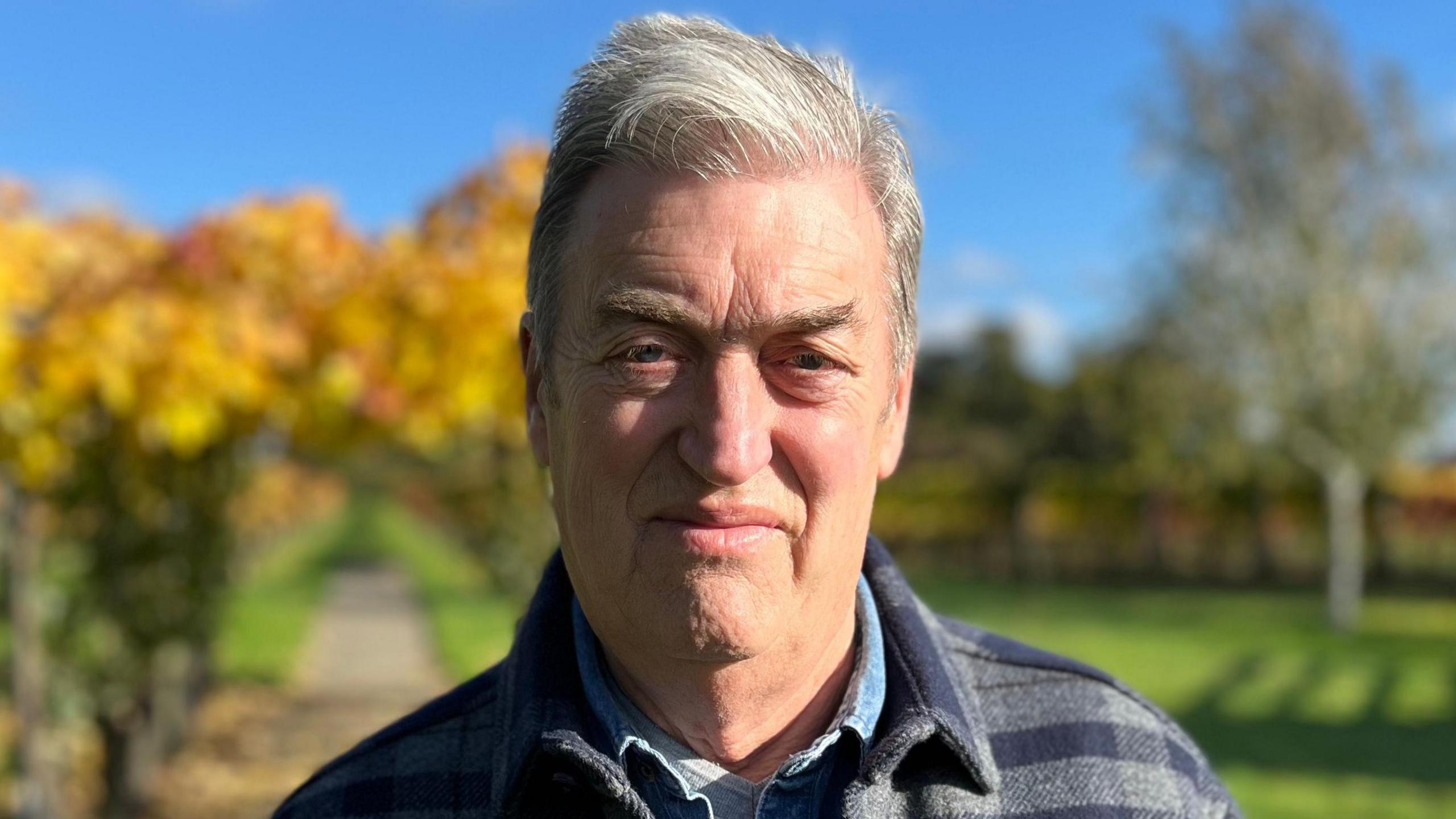 A picture of Les Garrett. He is standing in his vineyard and smiling at the camera. He has short grey hair and is wearing a blue and grey chequered coat on top of a blue shirt. 