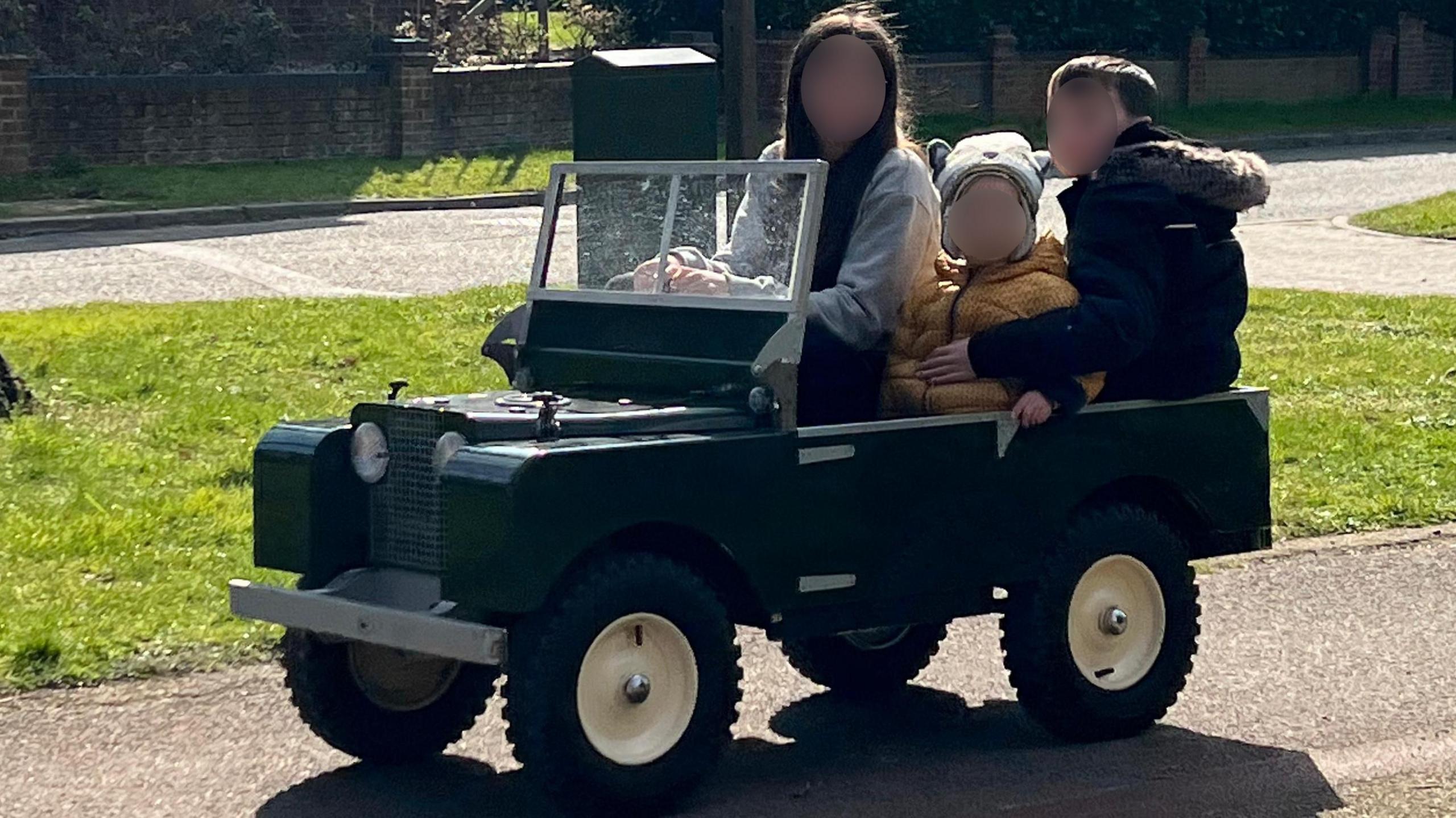 Three children are sitting in the mini-Land Rover, which is driving down a path on a housing estate. The children's faces have been blurred.