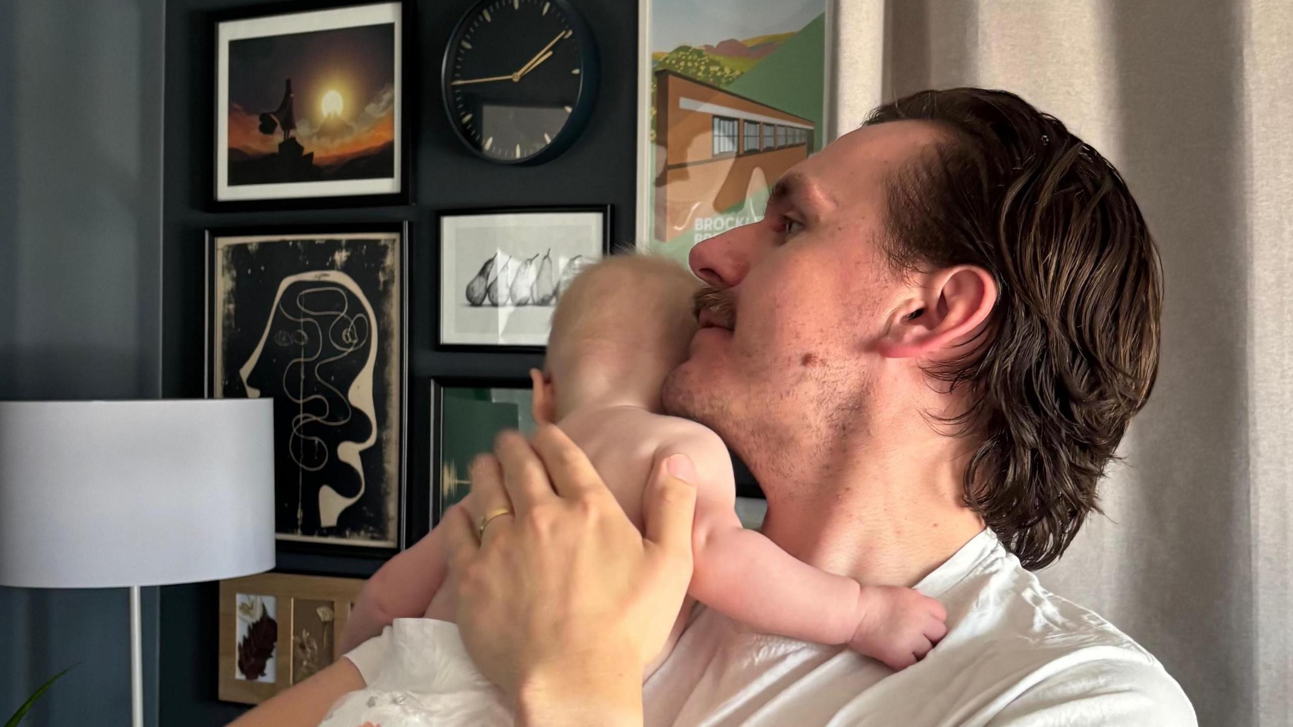 A man with brown hair and a moustache holds a baby to his shoulder