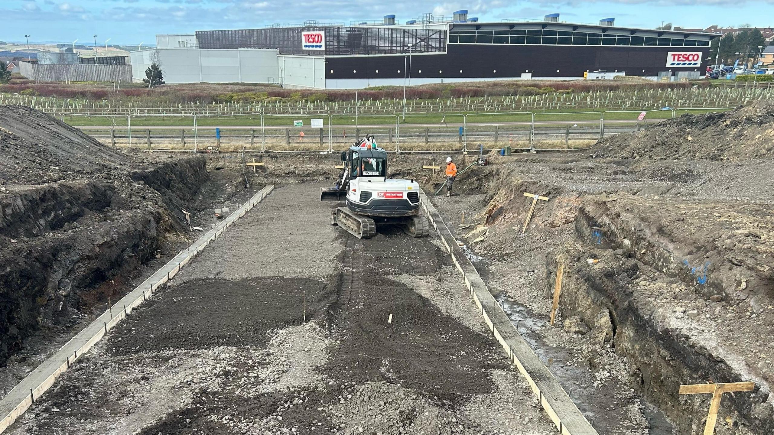 A bulldozer at the site of the Shotley Bridge Hospital. A section of the land has been dug out. There is a Tesco store behind the building site.