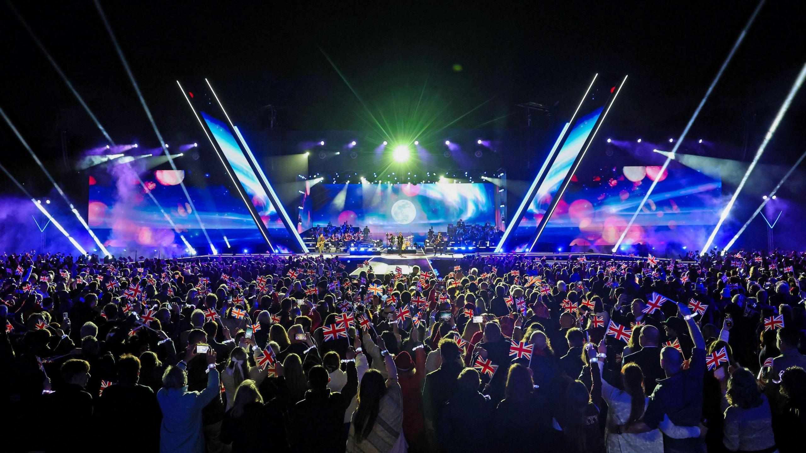 A large crowd stands in front of a stage. The stage has a pink and blue lights on its backdrop and an image of a full moon. Many people in the crowd are holding up small Union Jack flags.