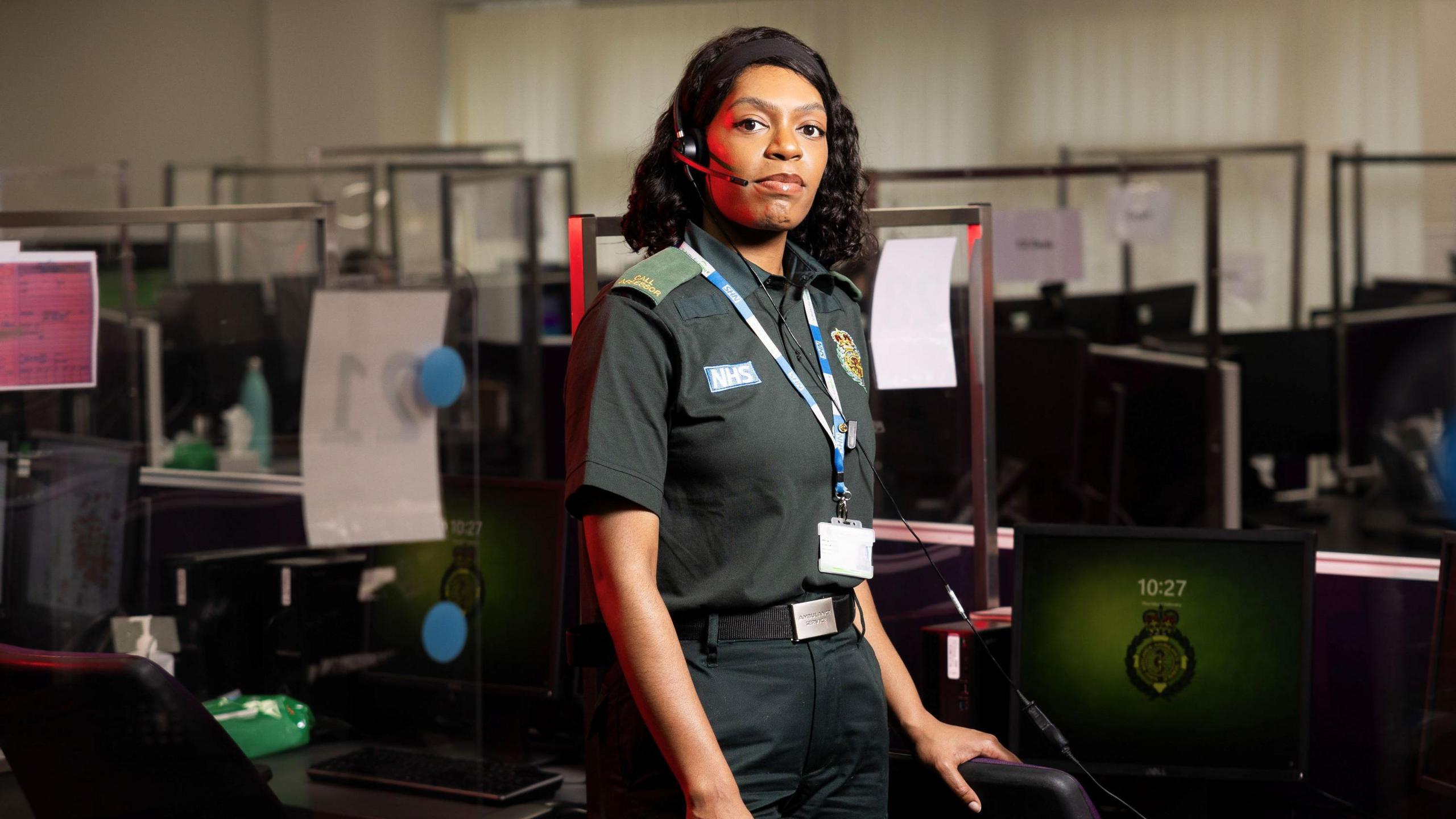 Laura Davis pictured in dark green West Midlands Ambulance Service uniform. She is looking at the camera and is wearing a headset and behind her are desks, computers and chairs. 