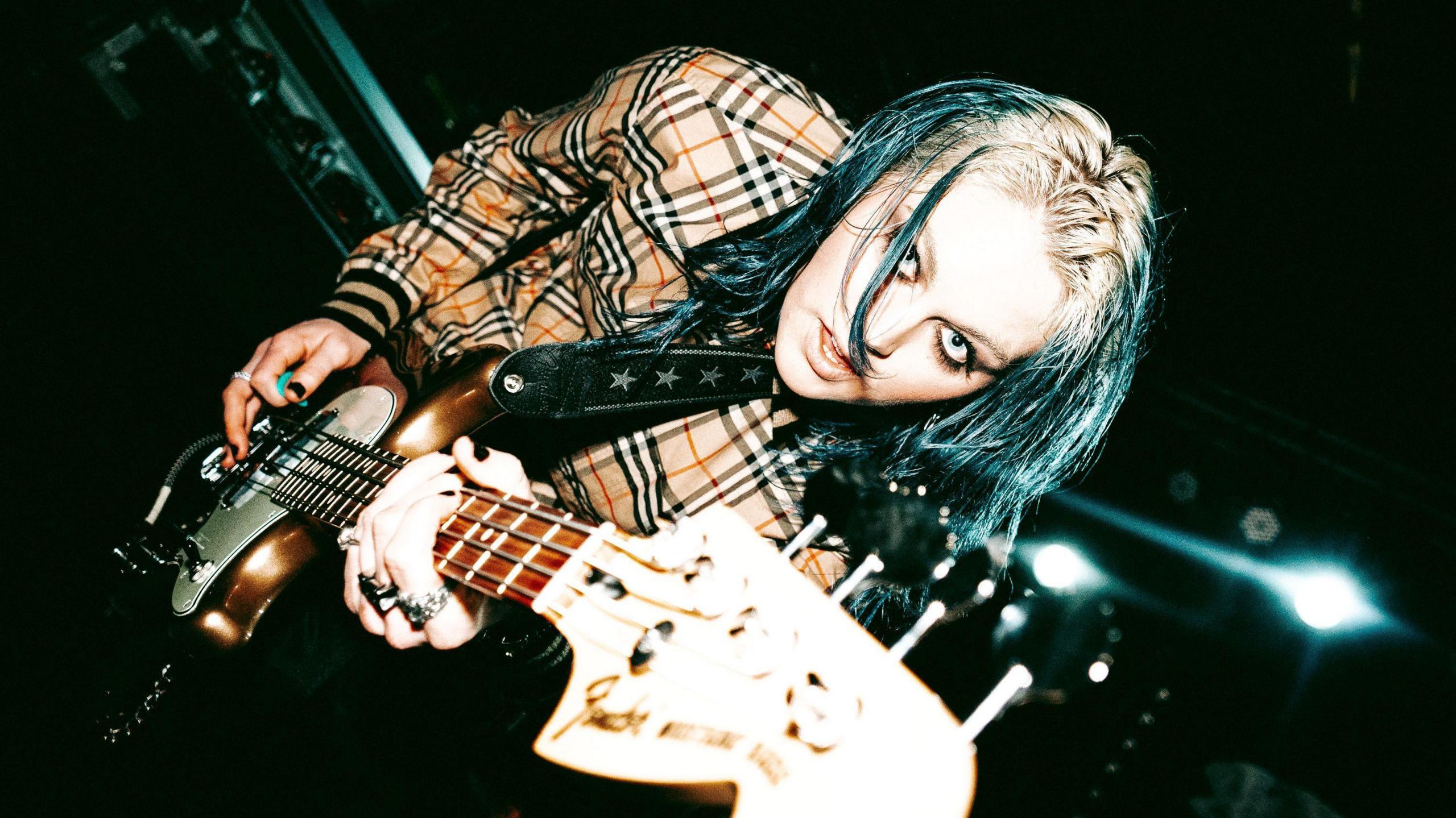 lleo, who has blonde and blue hair, wearing a Burberry shirt and playing a guitar. She has dark eye make up on and looks directly into the camera