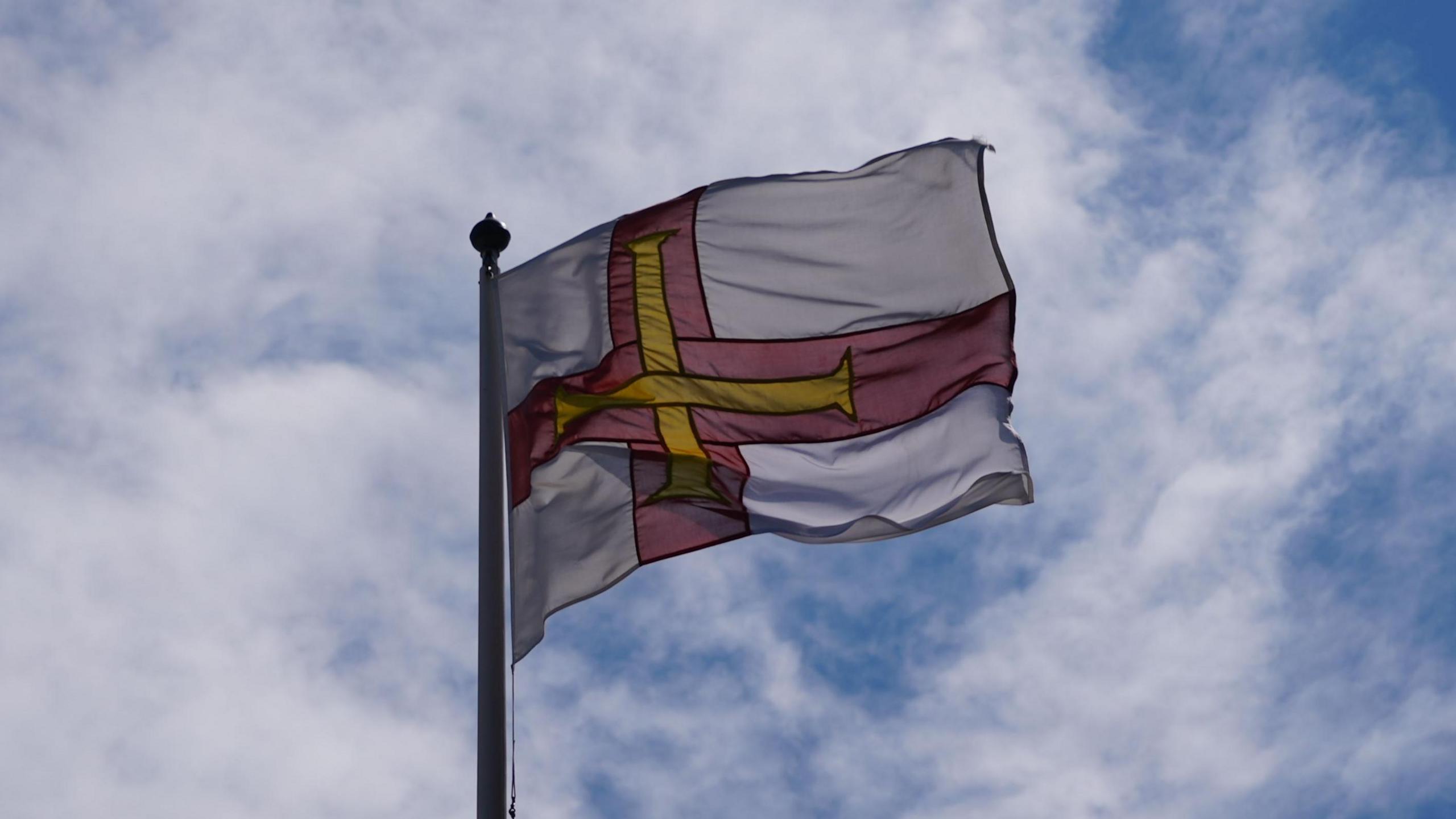 A Guernsey flag, which is a red cross with a yellow cross inside on a white background. It is flying in the wind and in the background is the sky and clouds. 