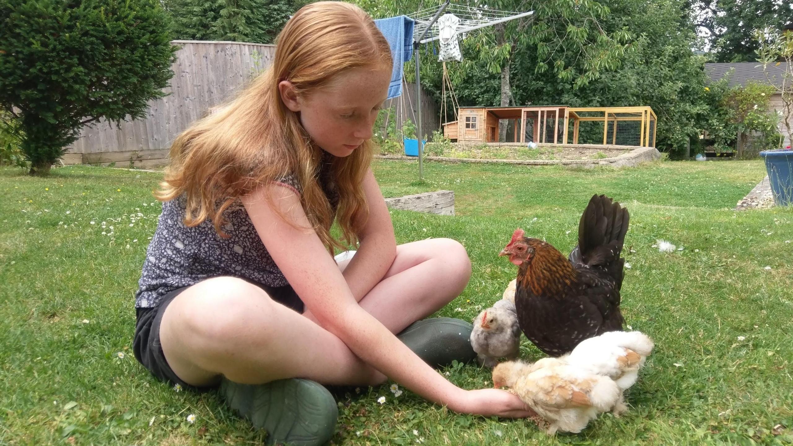 A picture of Ruth sitting on grass in what appears to be a back garden, with chickens eating food from her open hand. 