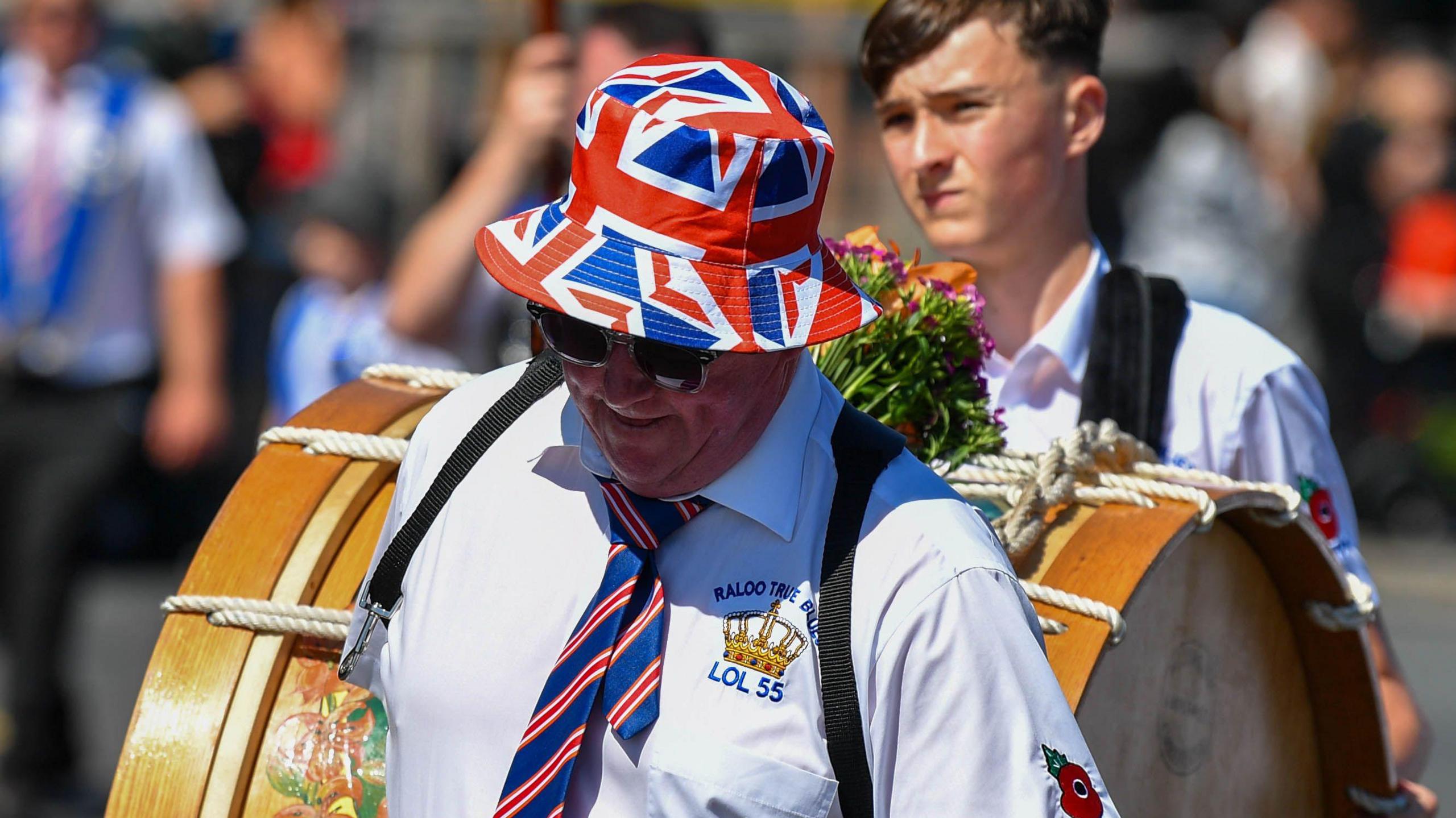 A man wearing a Union flag bucket hat holding the front of a drum. He is wearing a white shirt with a crown emblem on the left breast and a blue tie with red and white stripes.