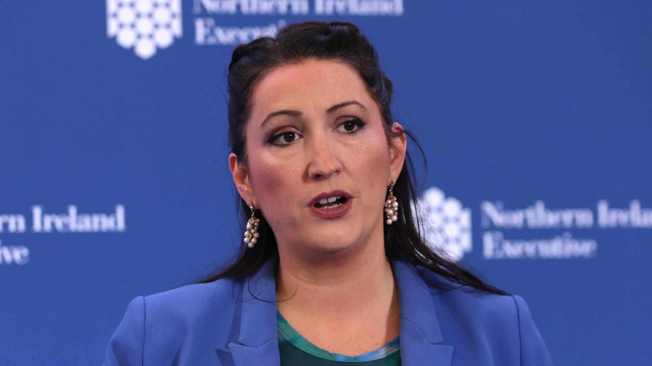 Emma Little-Pengelly. She has long dark hair partly tied back, wearing a blue blazer and pearl drop down earrings. A blue background is behind her with Northern Ireland Executive branding on it. She is standing in front of a microphone.