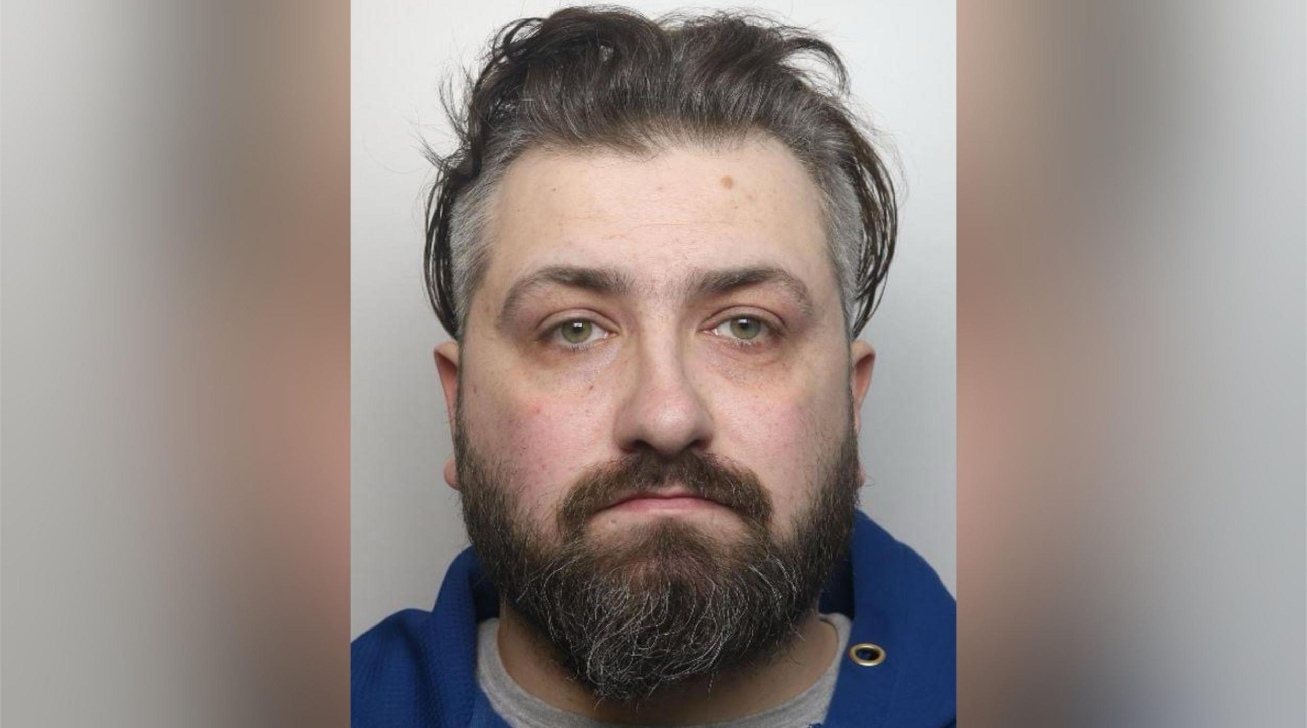 Alex Coleman with medium-length dark brown hair and beard, wearing a blue top in a police custody photograph.