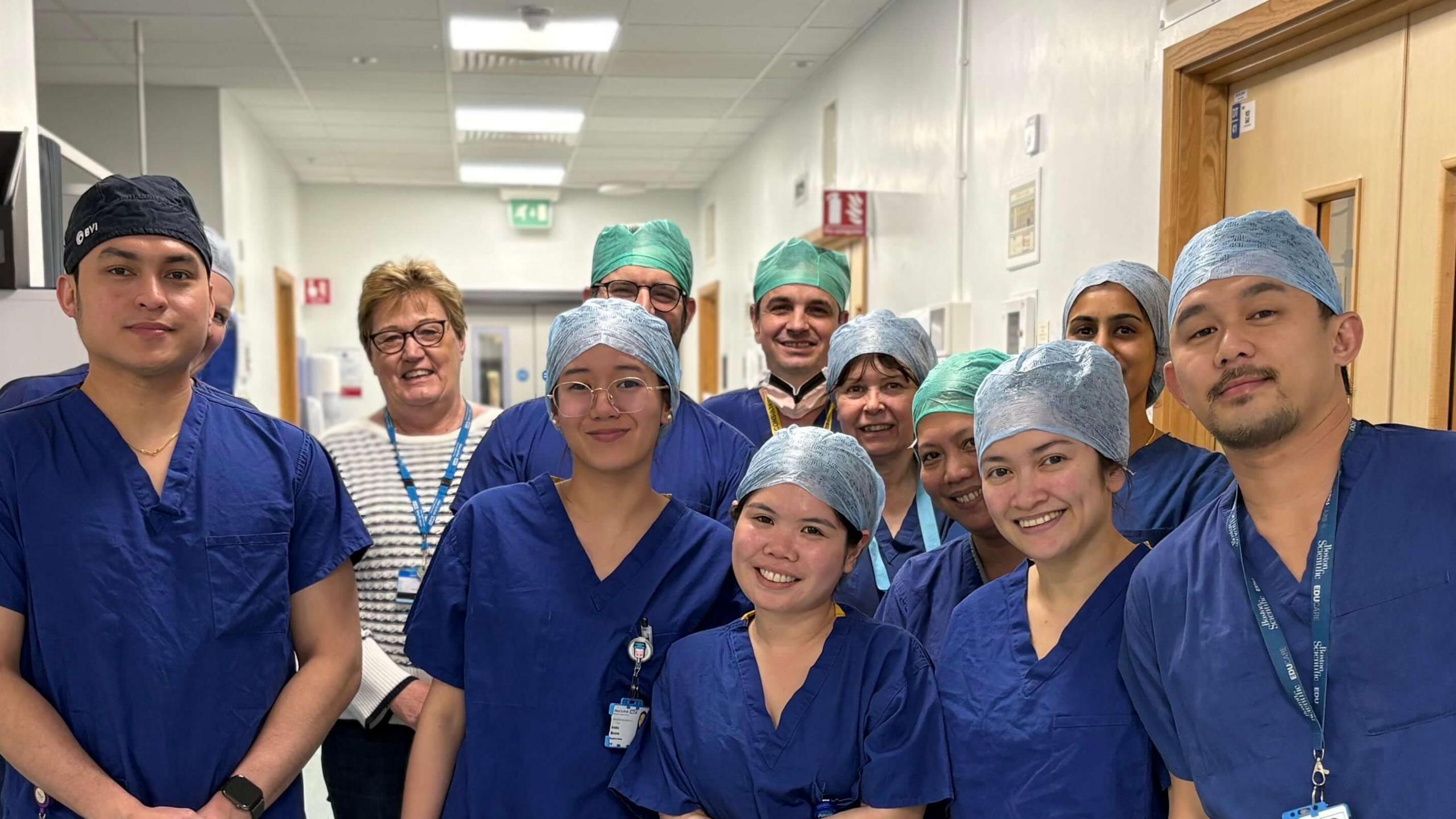 The West Suffolk surgeons stand together in a group wearing blue scrubs and hair covers. They smile at the camera.