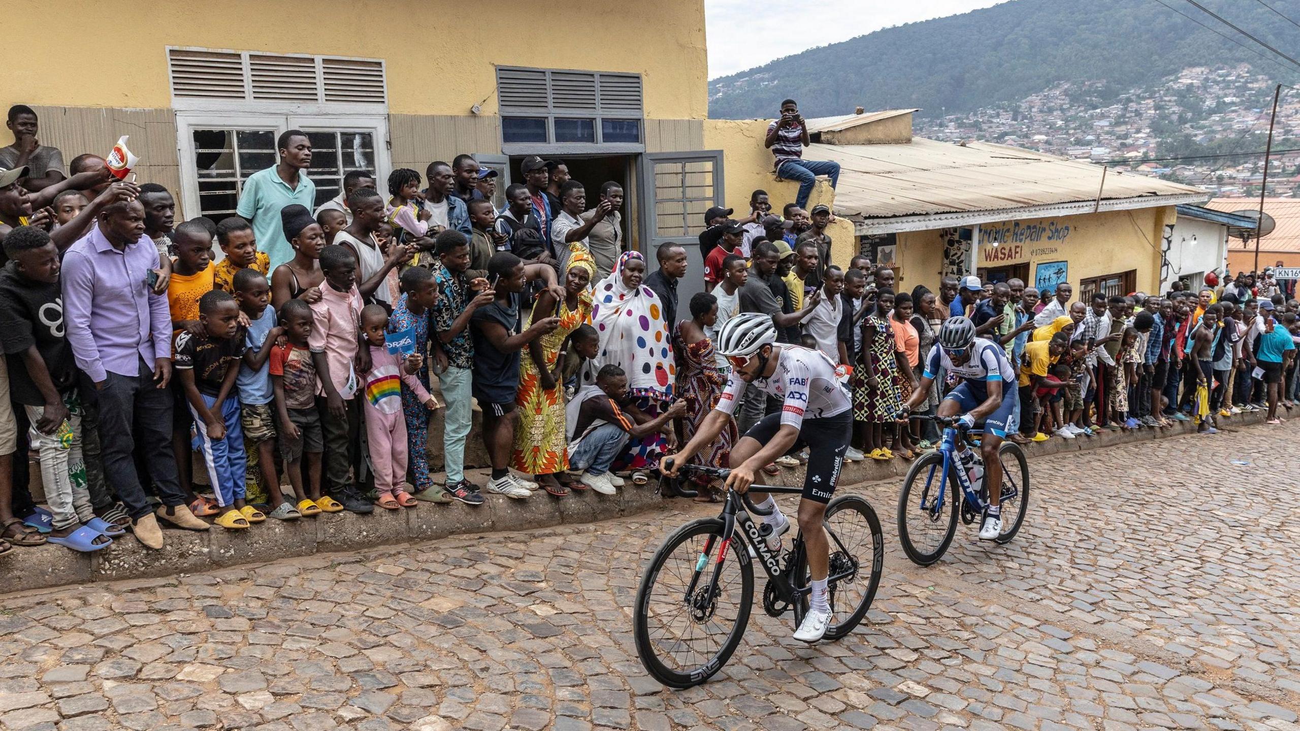 A crowd of almost 100 people line up in front of low-rise buildings on a section of a hilly, cobbled street in Kigali as two cyclists ride up the road in front of them. More of the city and a tree-covered mountain is seen in the distance