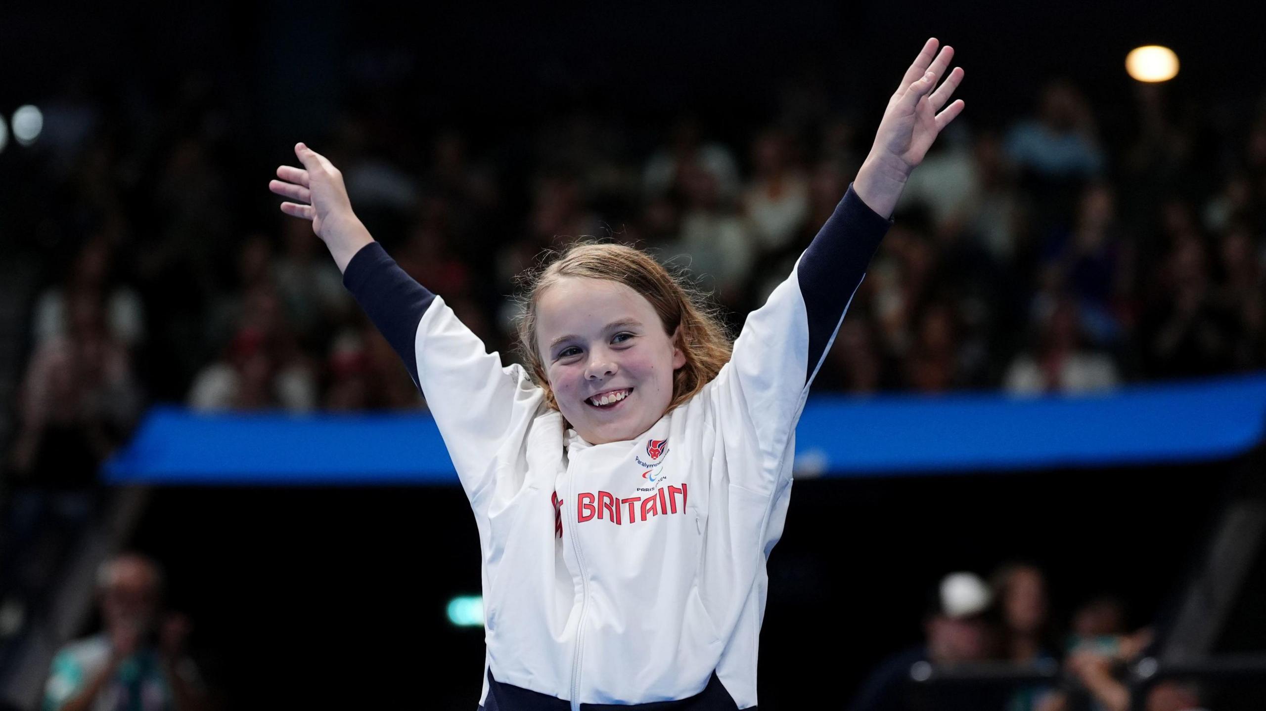 Iona Winnifrith holds both her arms in the air. She is wearing a white Great Britain paralympics uniform top and has a huge smile on her face. 