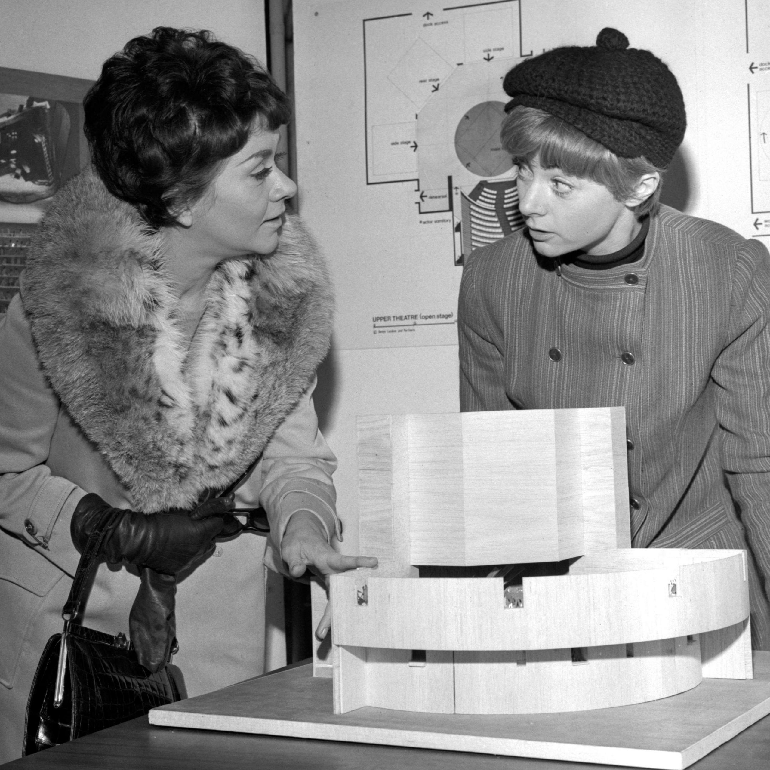 Joan Plowright and Geraldine McEwan talking and standing over a miniature model of the National Theatre, with architectural plans on the wall behind them