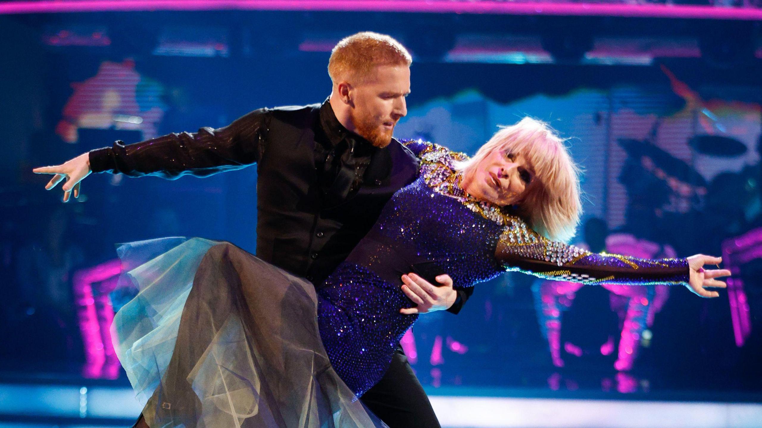 Neil holds Toyah as she leans away from him with her arm outstretched. Neil also holds his arm out. 