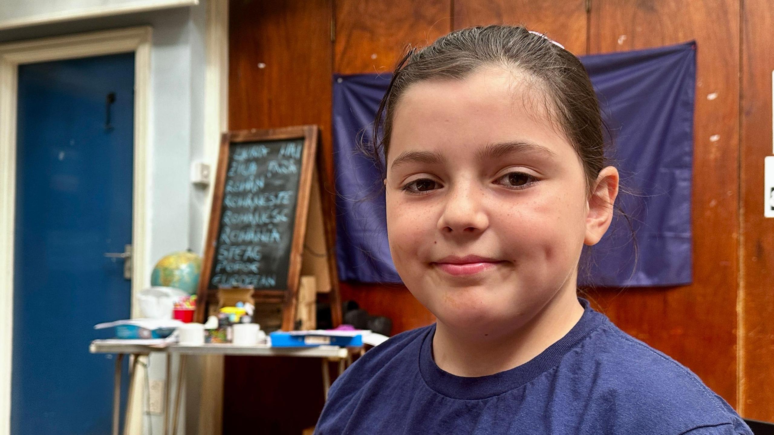 A nine-year-old girl is smiling and looking towards the camera. She has brown hair and a blue t-shirt.