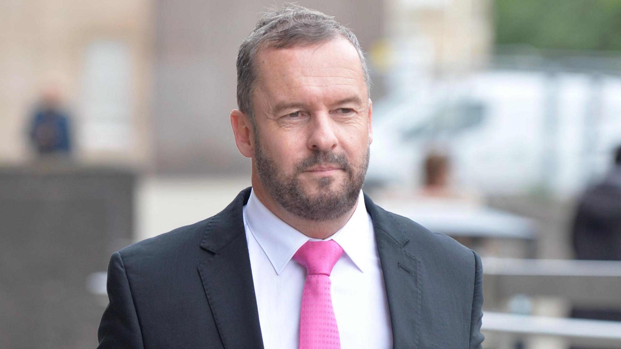 Andrew Polson, a man with thin grey/brown hair and a beard. He is walking down the street while wearing a dark suit and pink tie.