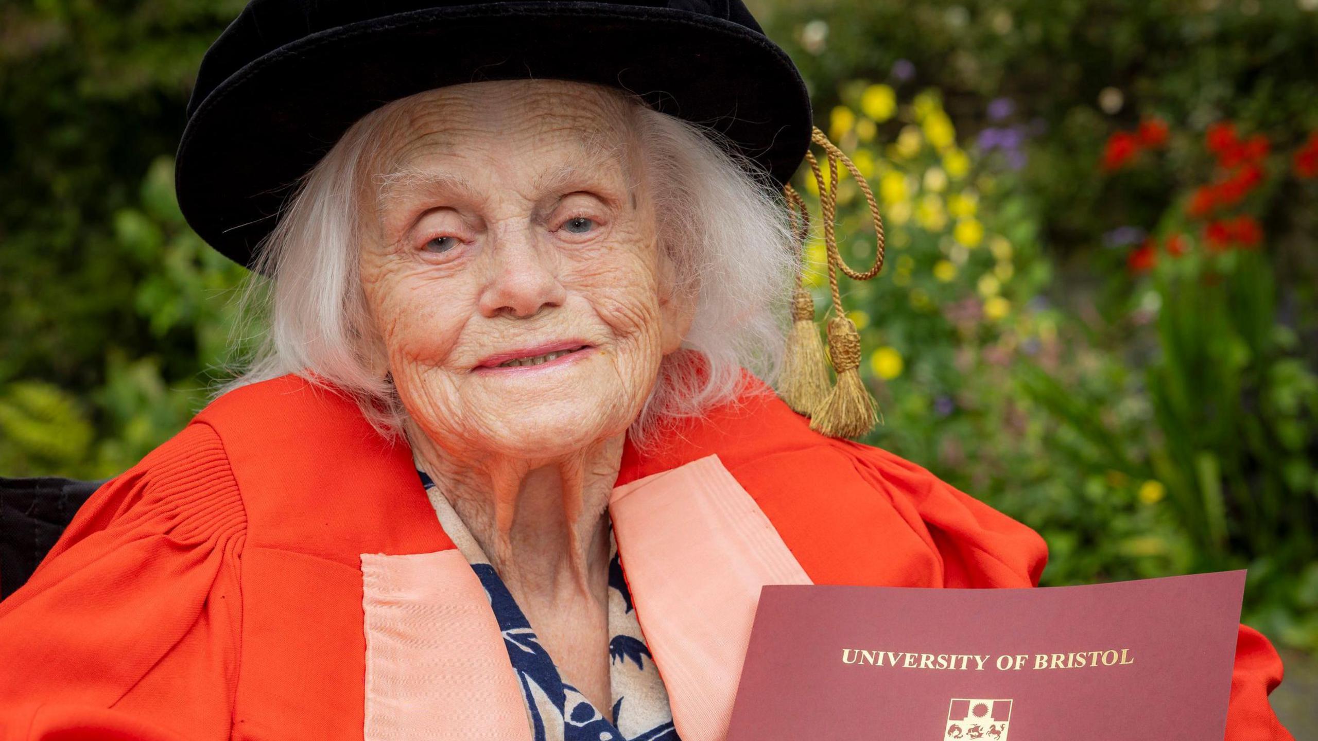 A woman sitting in  a wheelchair dressed in red graduation robes holds her doctorate certificate