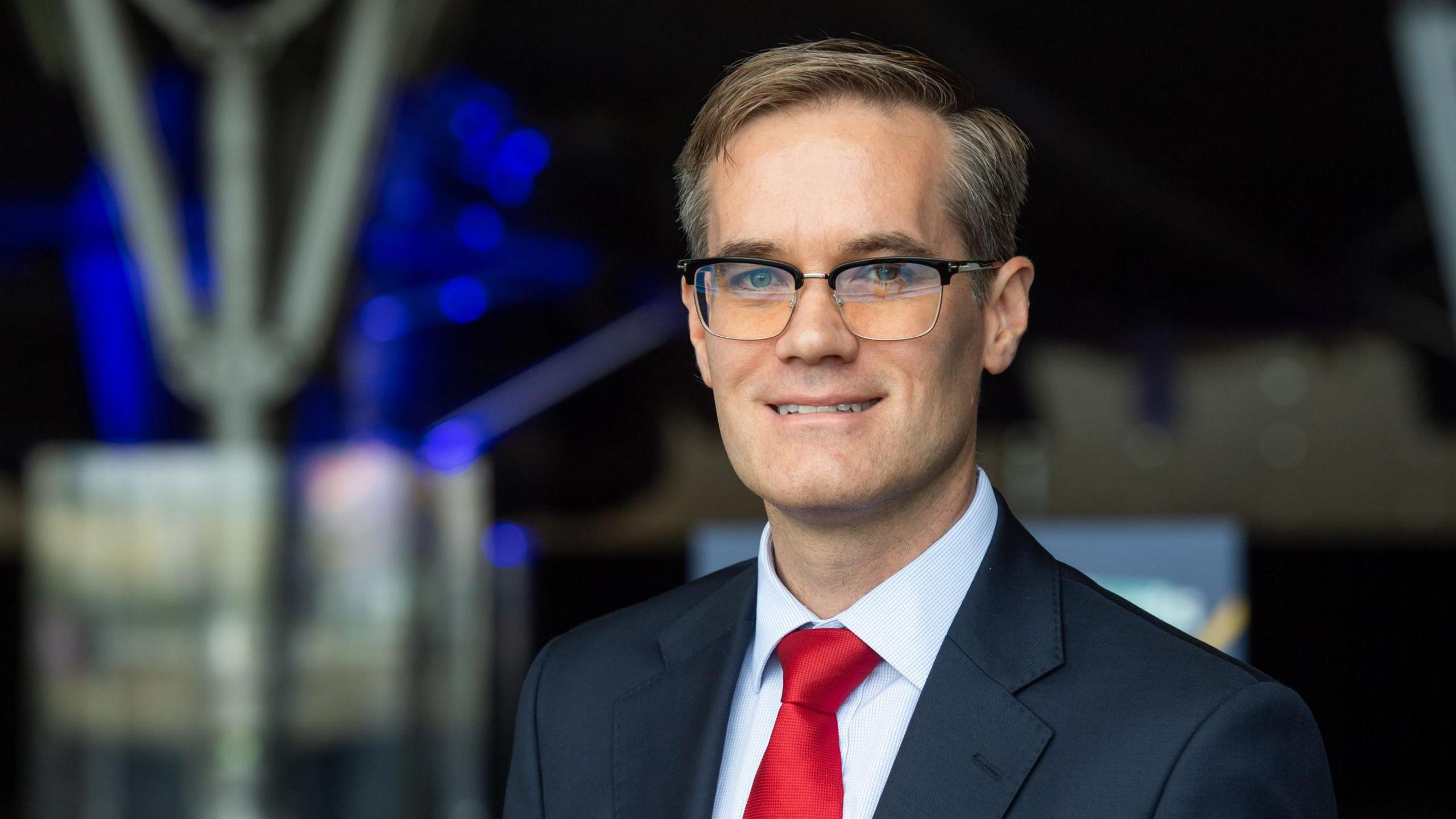 A man in a navy suit, red tie and glasses stands smiling at the camera.