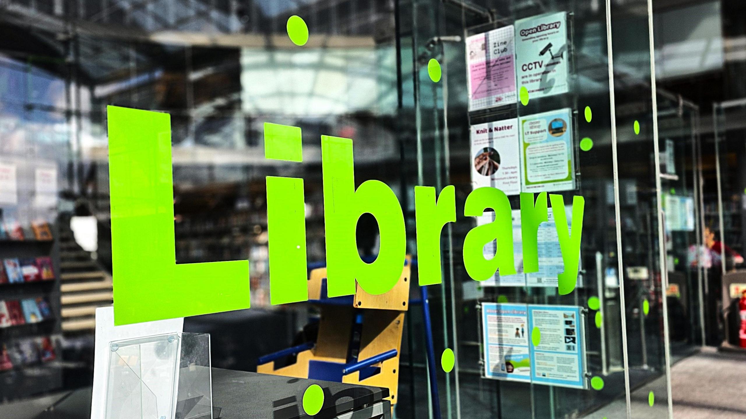 We can see a bright green 'Library' sign with letters stuck onto a glass panel. Behind it we can see the (out of focus) main Norwich library behind it.