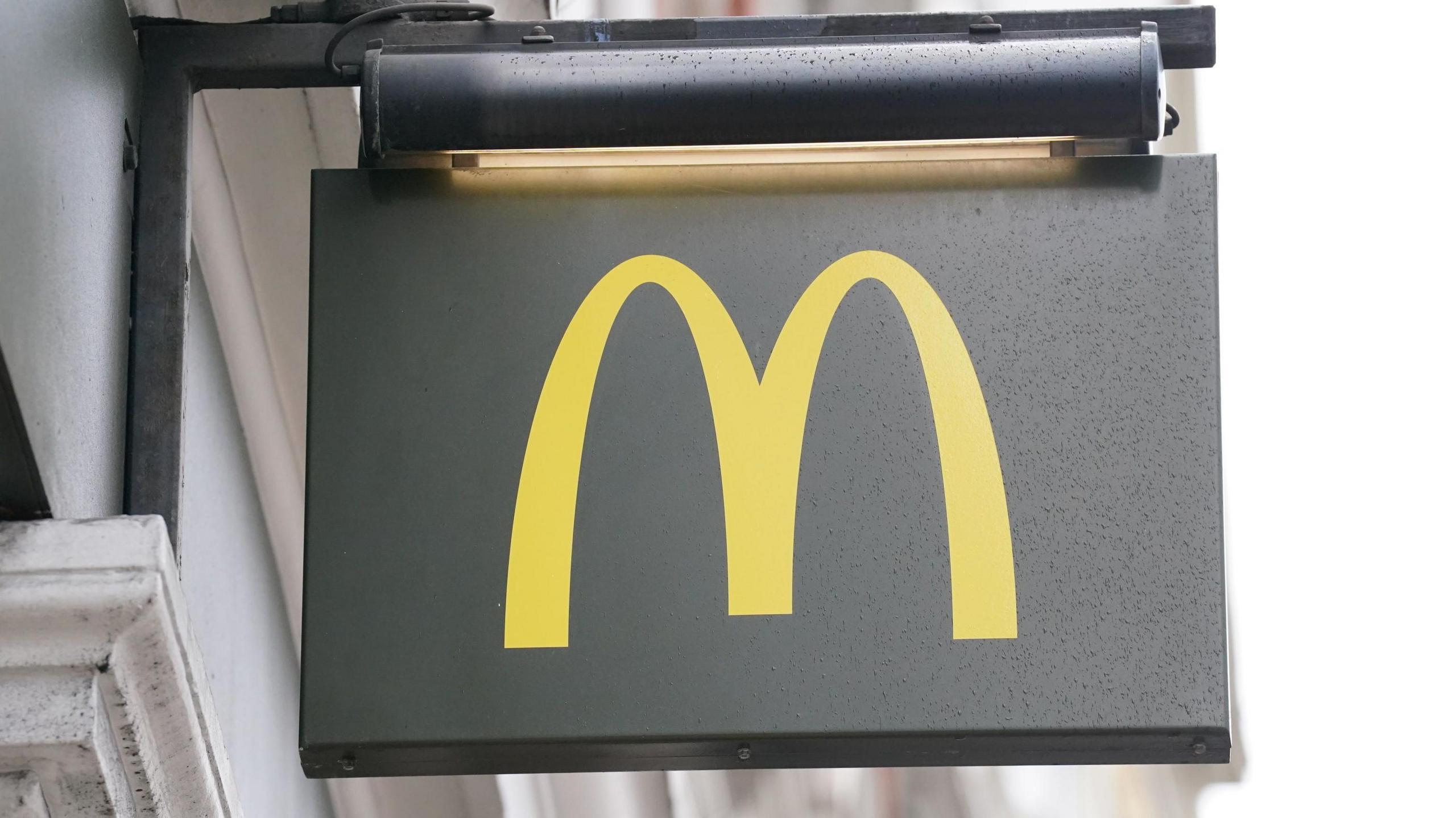 Signage for a branch of McDonald's. It is a dark grey sign with a yellow 'M' and it is it hanging on the wall of a white building.